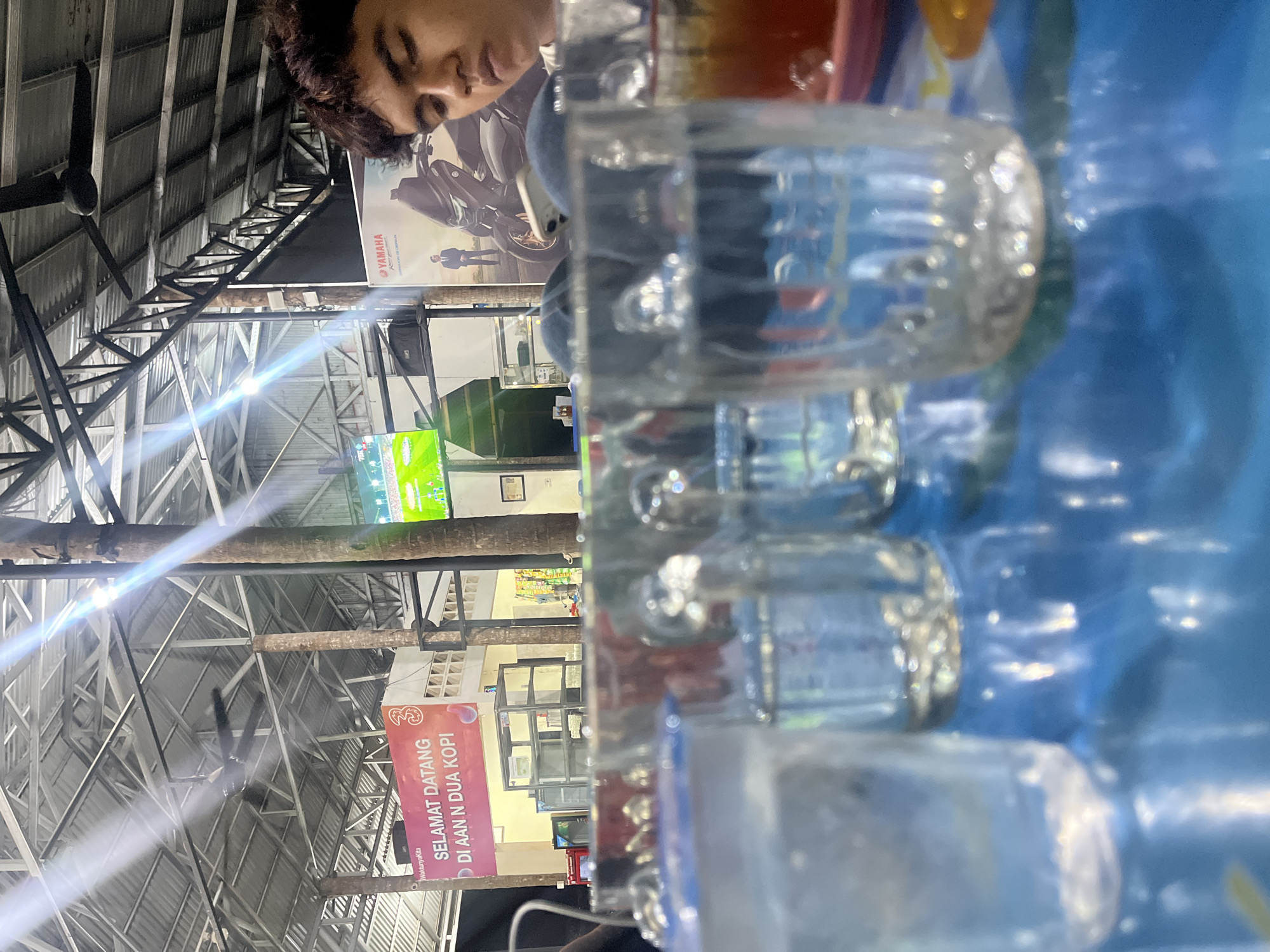 The image depicts an outdoor seating area, possibly a cafe or restaurant, in Banda Aceh, Indonesia. In the foreground, several glasses of water are on a blue table with a patterned surface. One glass, in the center, shows a reflection of the ceiling with lights and a fan. In the background, a man with curly dark hair is visible, partially obscured by the glasses. Behind him, a large poster features a Yamaha motorcycle and a man in a suit. Further in the background, a banner with red text that appears to say "SELAMAT DATANG DI AAN N DUA KOPI" is displayed, along with a television screen showing a soccer match. The environment suggests a casual, possibly evening setting, with overhead lighting and open-air structures. The presence of the Yamaha poster and the Indonesian text on the banner strongly suggests the location is within Indonesia, and "Aan N Dua Kopi" is likely the name of the establishment.