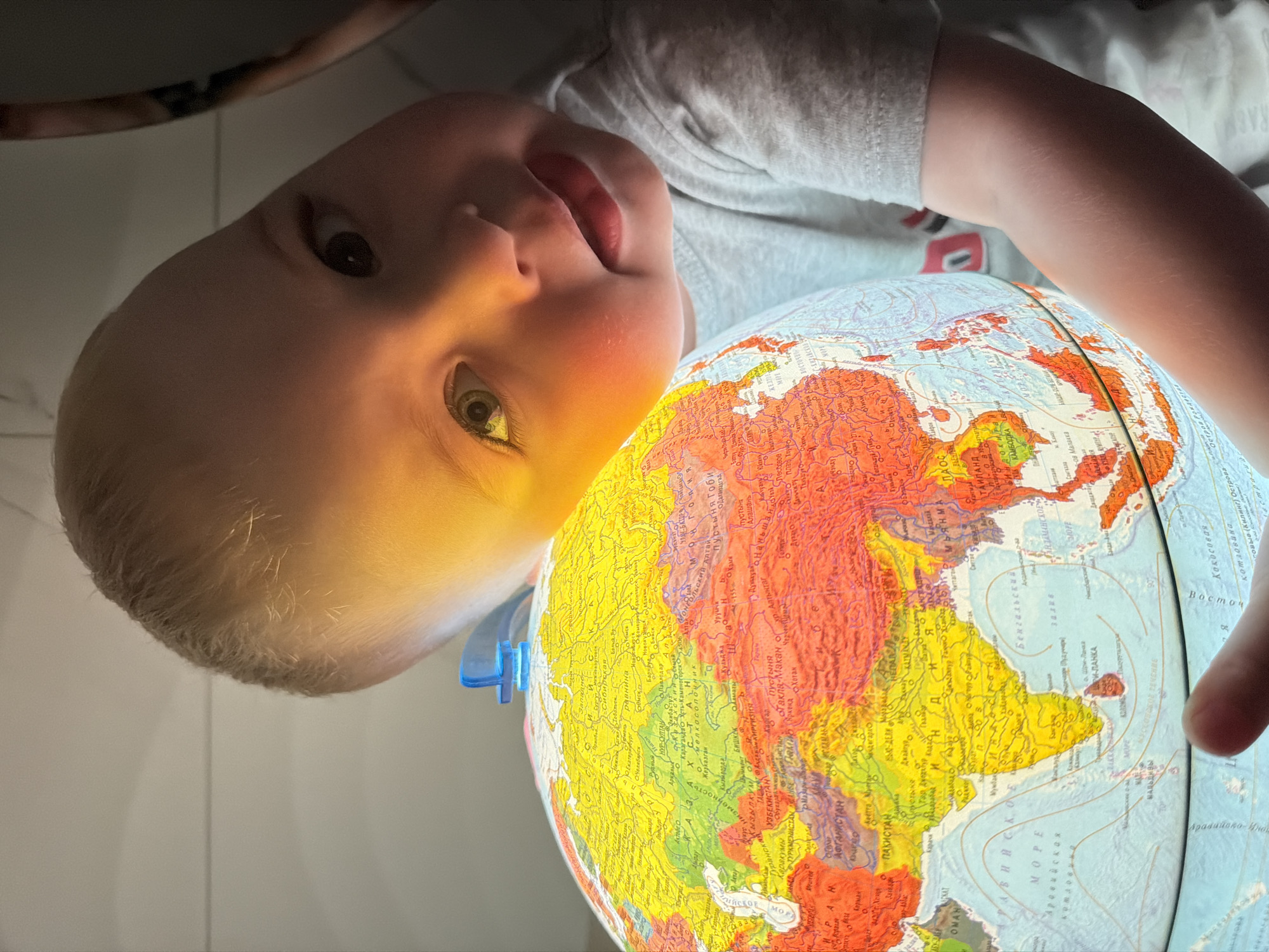 A child lies on the floor, positioned above and looking down at an illuminated globe. The globe dominates the scene, with its bright colors and visible text indicating geographical features. The child's face is close to the globe, with the light illuminating their skin and hair. The child is wearing a gray t-shirt. The background is a plain tile floor and hints of white walls or furniture. A blue plastic piece protrudes from the side of the globe. The globe is turned to show the continent of Asia.