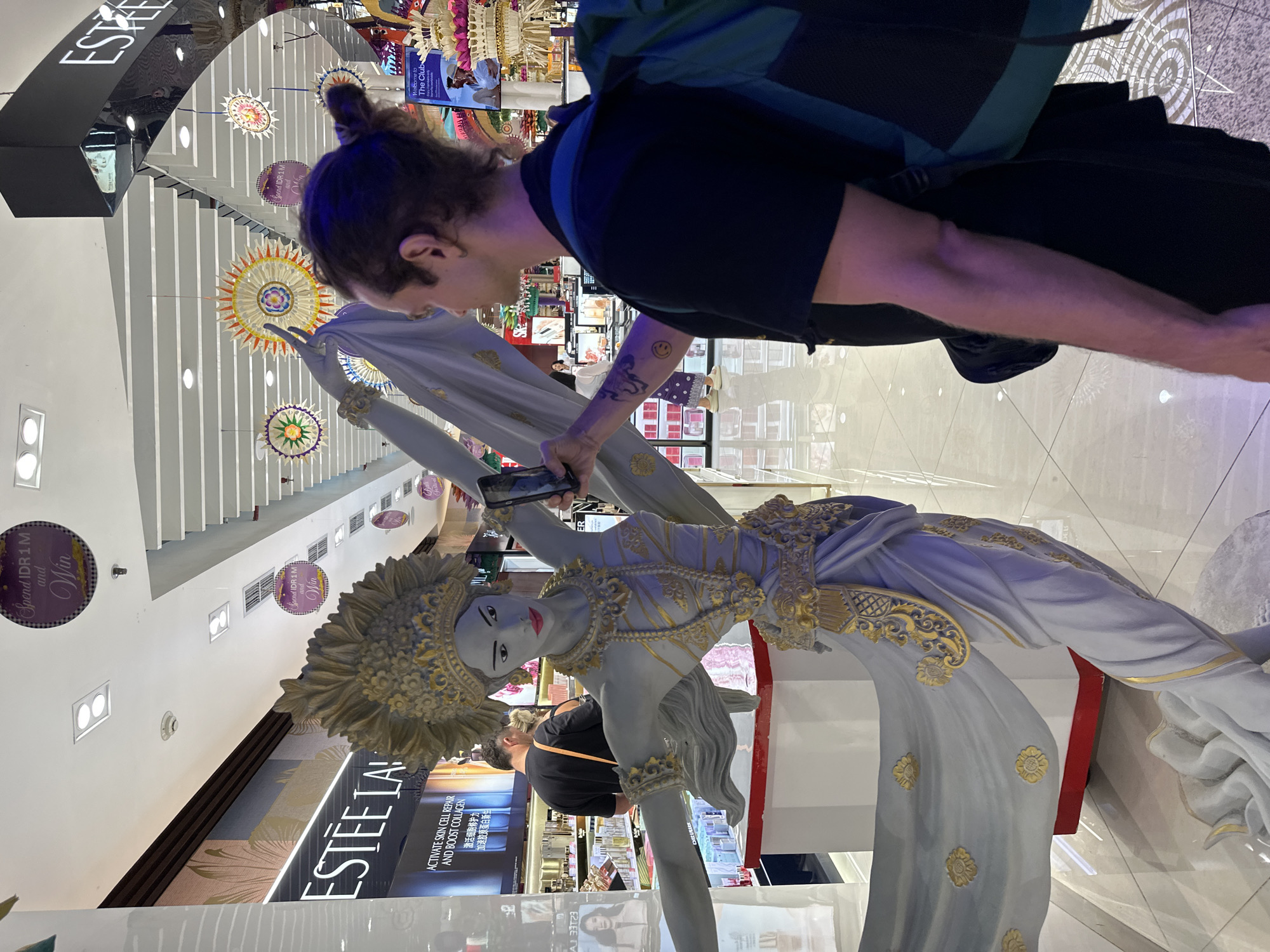 The image shows a light-skinned person with a man bun, wearing a black t-shirt and a backpack, taking a photo with a smartphone of a gray and gold statue of a woman in traditional clothing. The setting appears to be a high-end department store, possibly in the city of Kuta, Indonesia, given the "Spend IDR 1 M" signage and the Balinese-style decorations overhead. An Estee Lauder counter is visible in the background. The floor is light-colored and polished.