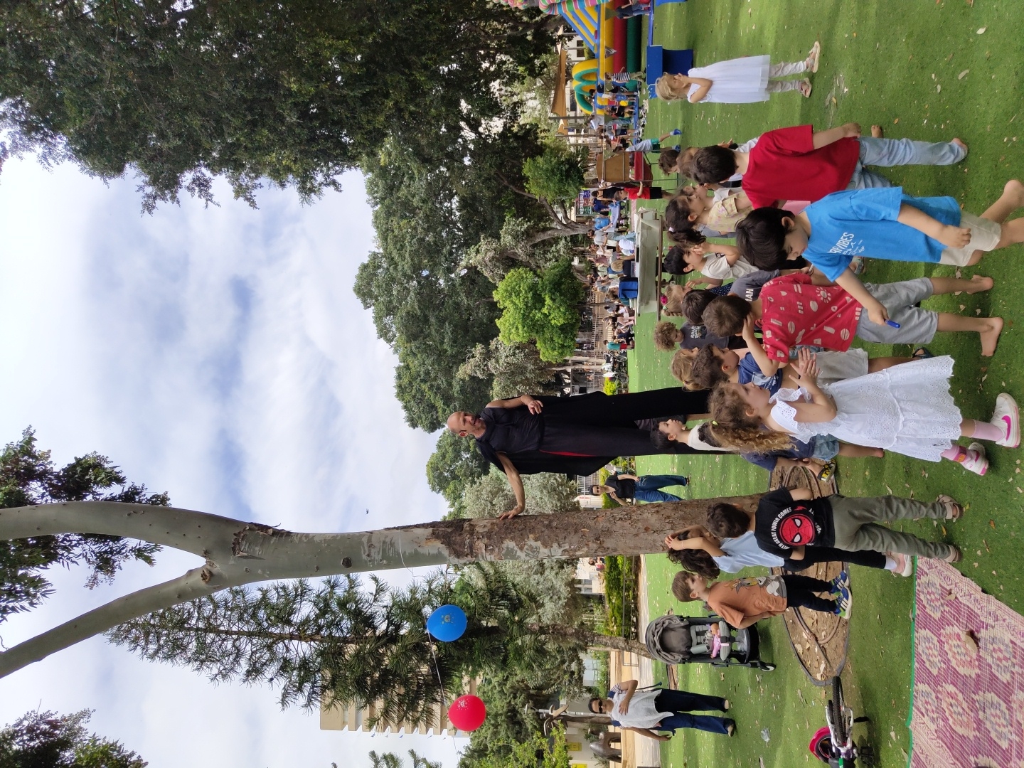 A group of children stands in line, watching a man in a black shirt and pants perched on a tree branch in what appears to be a park setting in Tel Aviv, Israel. Behind the children, a large crowd is gathered. The background also features green trees, buildings, and a bouncy castle. A few blue and red balloons are tied to a nearby tree.
