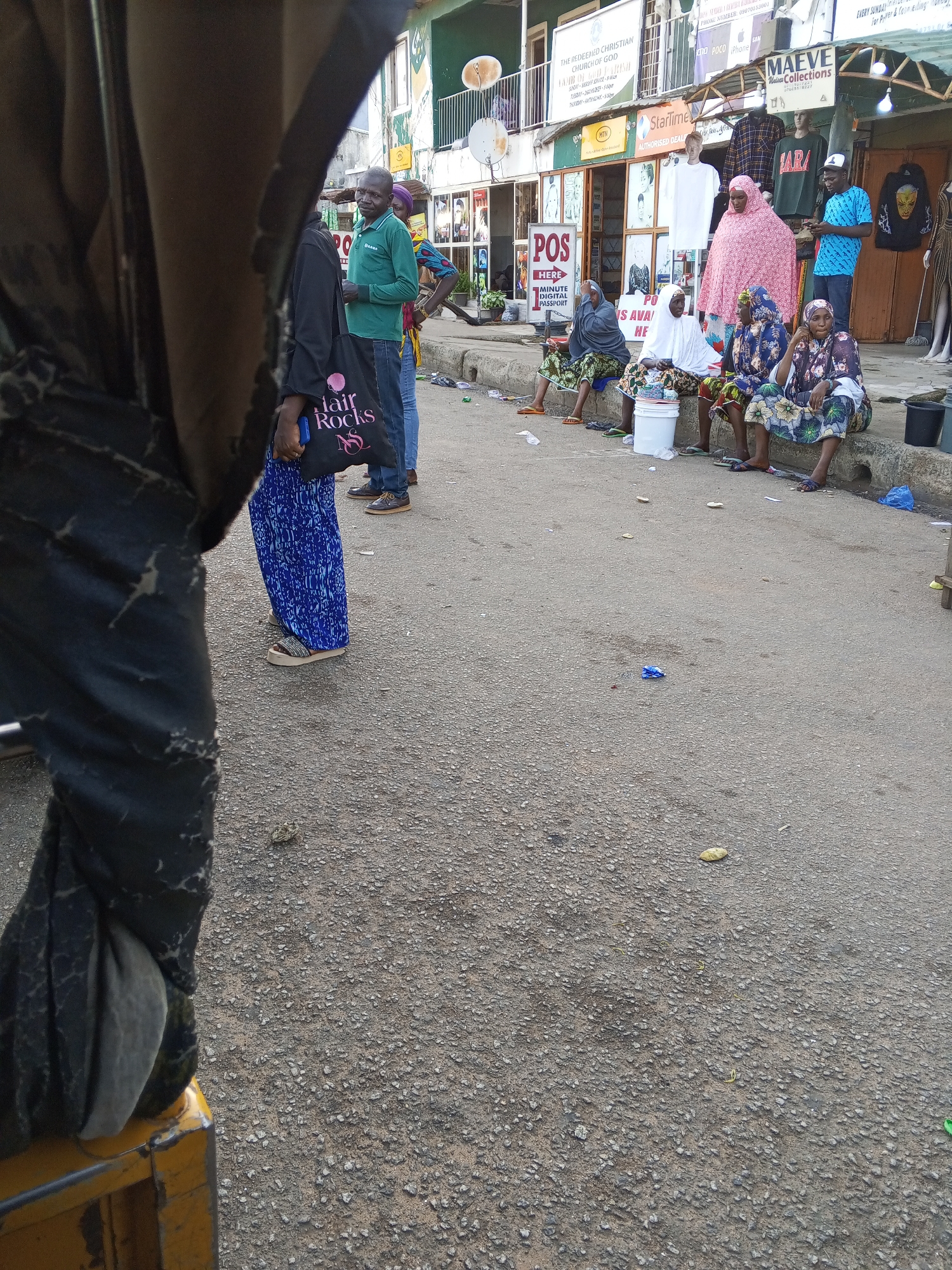 A street scene in Jos, Nigeria, captures a group of individuals in front of commercial establishments. In the foreground, the side of a yellow, motorized rickshaw is visible, partially obscuring a person wearing dark pants and a black garment. A black tote bag with "Hair Rocks" printed on it is held by a person whose lower body is visible, wearing blue patterned trousers and sandals.
Further back, a man in a green long-sleeved shirt and blue jeans stands talking to a person obscured from view. Several women are seated on a concrete curb, some wearing hijabs and patterned dresses. One woman in a pink hijab stands near a display of mannequins. Behind the group, shops are visible with signs including "MAEVE Collections" and "POS." The ground is paved with asphalt and appears somewhat littered with debris. The overall impression is of a busy marketplace or commercial street.