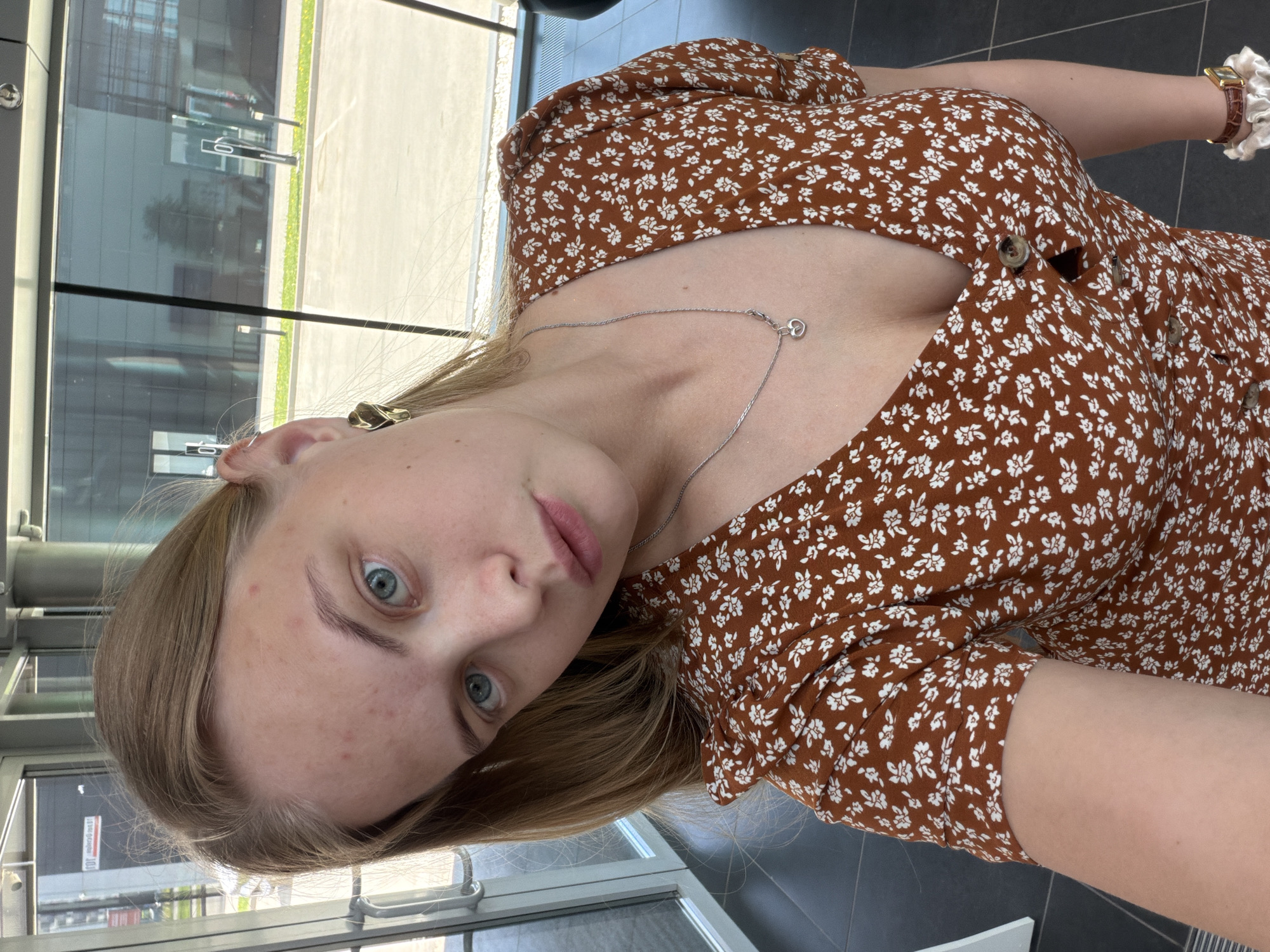 A young woman with blonde hair and blue eyes is taking a selfie. She is wearing a brown, floral-patterned dress with buttons down the front. A silver necklace is visible on her chest, and she has a watch with a white scrunchie on her wrist. The background shows an interior space with large windows reflecting the outside. Based on the reflections and style of architecture, it is highly possible that this photo was taken in a building located in Izhevsk, Russia.