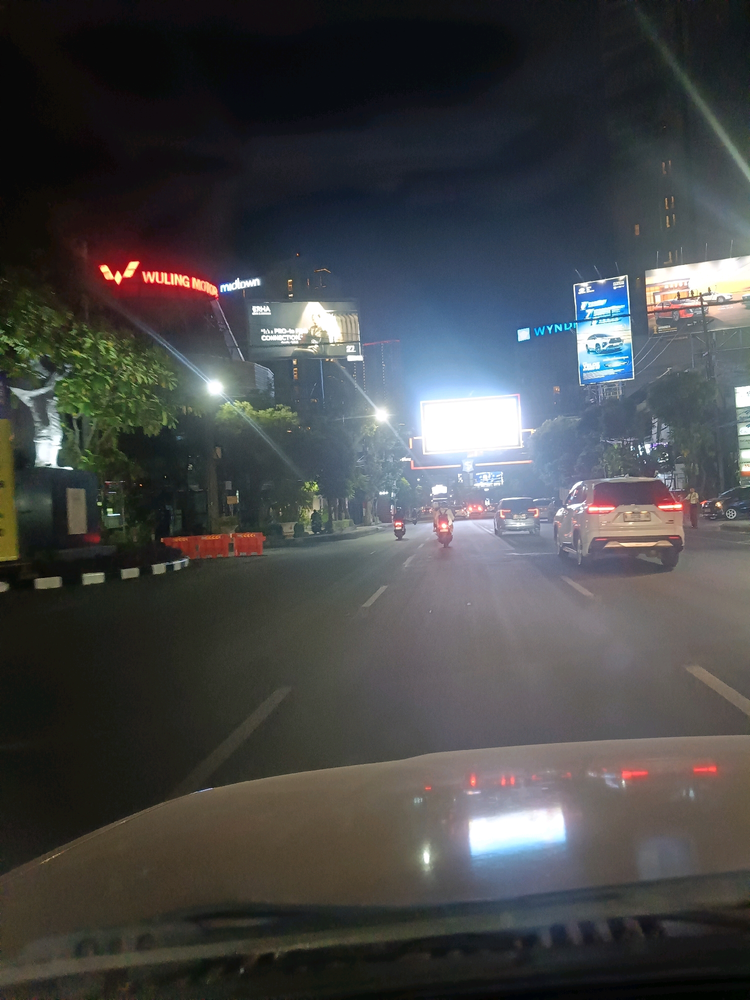 The image depicts a street scene at night in Surabaya, Indonesia. A prominent red neon sign with the text "WULING MOTORS midtown" is visible on the left side of the frame. To the right, a large billboard displays advertisements for cars, with the "WYNDHAM" logo also present. The street is lined with trees and illuminated by streetlights. Several vehicles, including cars and motorcycles, are driving on the road. In the foreground, the dashboard of a vehicle is visible, reflecting streetlights and the interior of the car. The overall atmosphere suggests a bustling urban environment at night, likely a commercial or entertainment district.