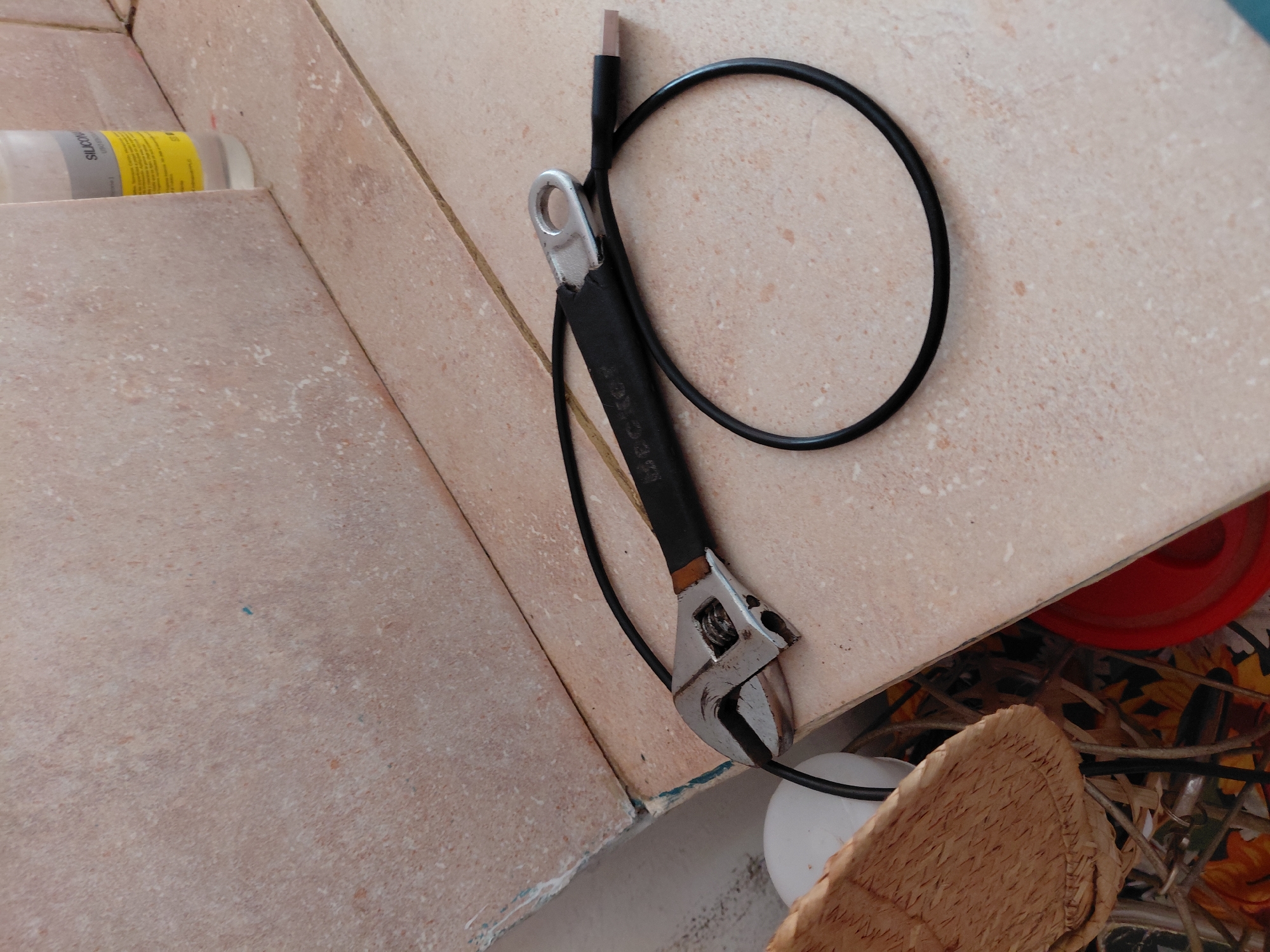 The image captures a collection of objects resting on and around a light-colored tiled surface, likely in a utility area such as a bathroom, kitchen, or workshop in Santa Ana, Venezuela.
The primary subjects are an adjustable wrench (often called a monkey wrench) and a black cable. The wrench has a metallic head and a black handle, with "DECKER" faintly visible on the handle, suggesting a brand like Black & Decker. The cable, which appears to be a charging or data cable, has a USB-C type connector visible at one end and is coiled in a loop. It passes through the circular opening at the end of the wrench's handle.
In the upper left, a white plastic bottle with a yellow label is partially visible, resting on a lower tiled surface. The text "SILICON" is clearly legible on the label, indicating it is likely a silicone sealant or caulk product.
The tiles are a mottled light beige or pinkish-brown, suggesting a speckled or textured finish. They form a corner, with some chipped edges and residue (possibly old sealant or paint) visible along the grout lines and at the bottom edge.
Below the main tiled surface, on the right side, other items are partially visible: a red plastic object (possibly a bowl or bucket), a woven straw-like material (perhaps a mat or part of a basket), and a patterned fabric featuring yellow and orange shapes, possibly sunflowers.
The lighting is bright and even, suggesting daytime or a well-lit indoor environment. There is no specific activity taking place; the objects are static, implying they have been set down or stored. The presence of a wrench and silicone sealant suggests potential for repair, maintenance, or construction work in the area. No people are visible, and no specific weather conditions can be discerned from the image.
