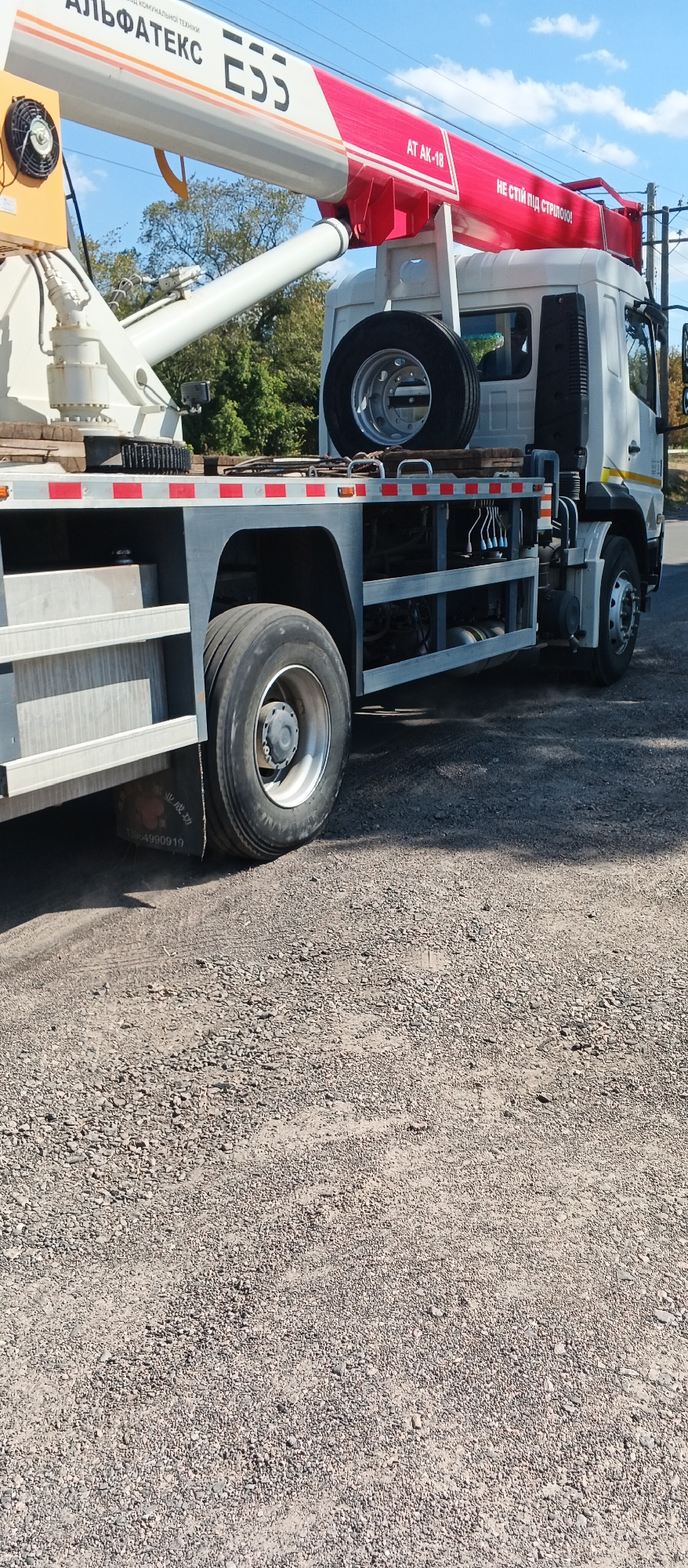 An outdoor, low-angle medium shot captures a white truck with a red crane arm. The truck has a spare tire mounted vertically on its side and a black fuel tank. The crane arm is labeled with "АТ АК-18" and Cyrillic text. The truck is parked on a gravel surface, with trees visible in the background. The scene appears to be an industrial area or roadside in Smila, Ukraine, under a partly cloudy sky.