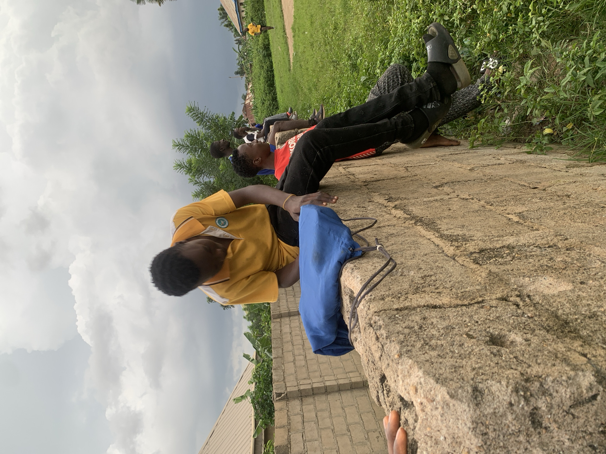 In Sabon Tasha, Nigeria, under a cloudy sky, several young men are gathered outdoors. The foreground features a rough, concrete wall where a young man in a yellow polo shirt and black pants is seated, looking down. A blue bag is placed on the wall beside him. To his right, another young man in a red shirt and black pants is also seated on the wall, looking to the side. Behind them, two other young men are seated further down the wall. In the background, there is lush green grass, trees, and buildings. A lone figure in a yellow shirt is visible running in the distance. The overall scene depicts a casual gathering in a semi-urban environment.