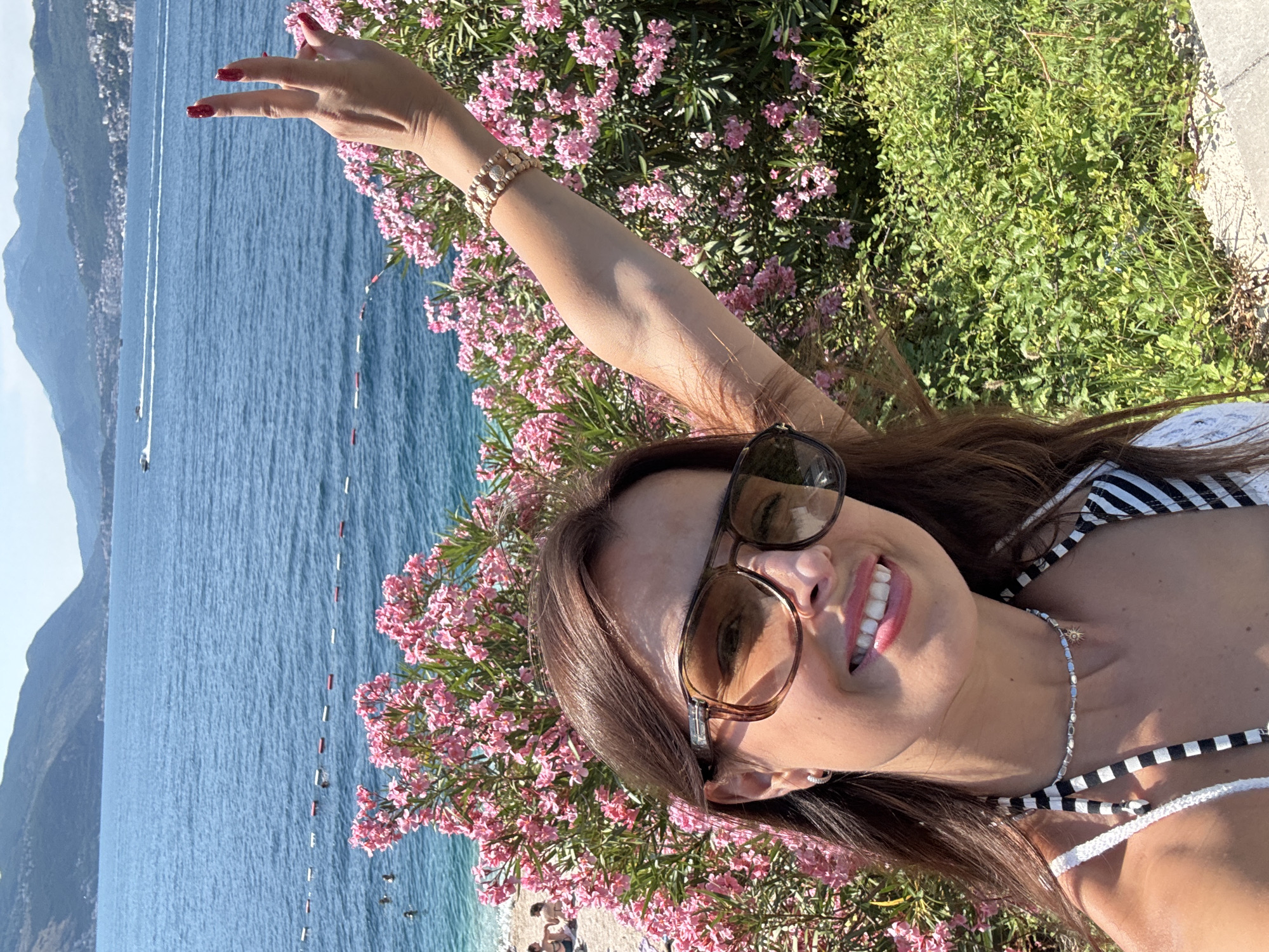 A medium shot captures a woman with brown hair and sunglasses taking a selfie. She is wearing a black and white striped bikini top and a necklace with a sun pendant. Her right arm is raised, and she is making a peace sign with her fingers. Behind her, pink oleander flowers bloom abundantly, and beyond that, the blue sea stretches to distant hills under a clear sky. Several buoys mark a swimming area in the sea, and a boat can be seen in the distance. The woman is smiling, and the overall scene suggests a sunny day at a coastal location.