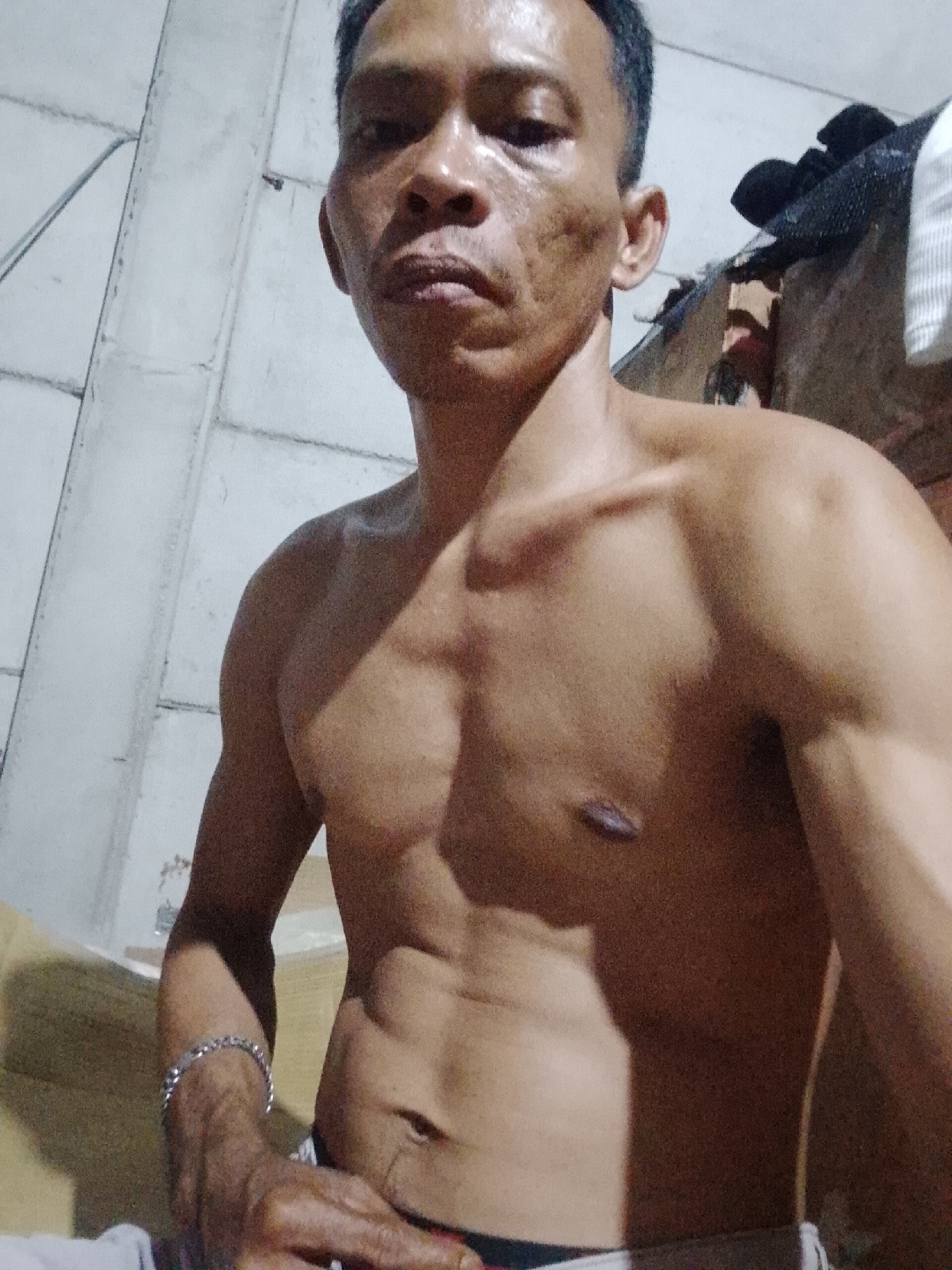 The image depicts a shirtless man with a muscular build. He has short, dark hair and appears to be indoors. He is wearing a bracelet on his left wrist. The background includes a wall with visible seams. The location, Bukit Ketok, Indonesia, provides geographical context but is not directly visible in the scene.