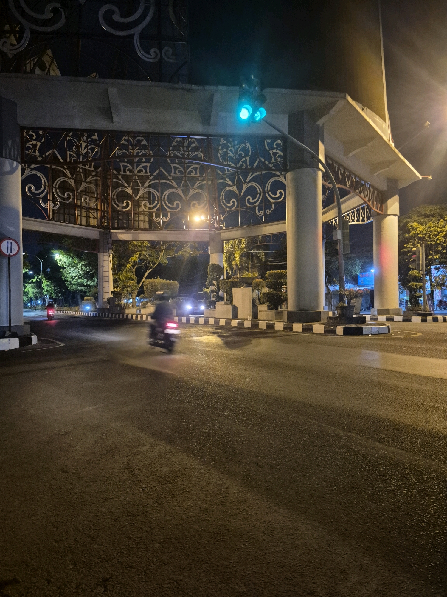 The image depicts a nighttime scene at a traffic intersection in Surakarta, Indonesia. A traffic light overhead is illuminated green. The intersection features a large, ornate gateway structure supported by thick pillars. The road surface appears wet, reflecting the ambient light from street lamps and vehicle headlights. In the foreground, a motorcycle rider is blurred due to motion, with red taillights visible. Another motorcycle, also blurred, is further down the road on the left, with its red taillight also visible. Trees and decorative bushes line the sides of the road. The overall impression is of a moderately busy intersection at night, possibly after rain.
