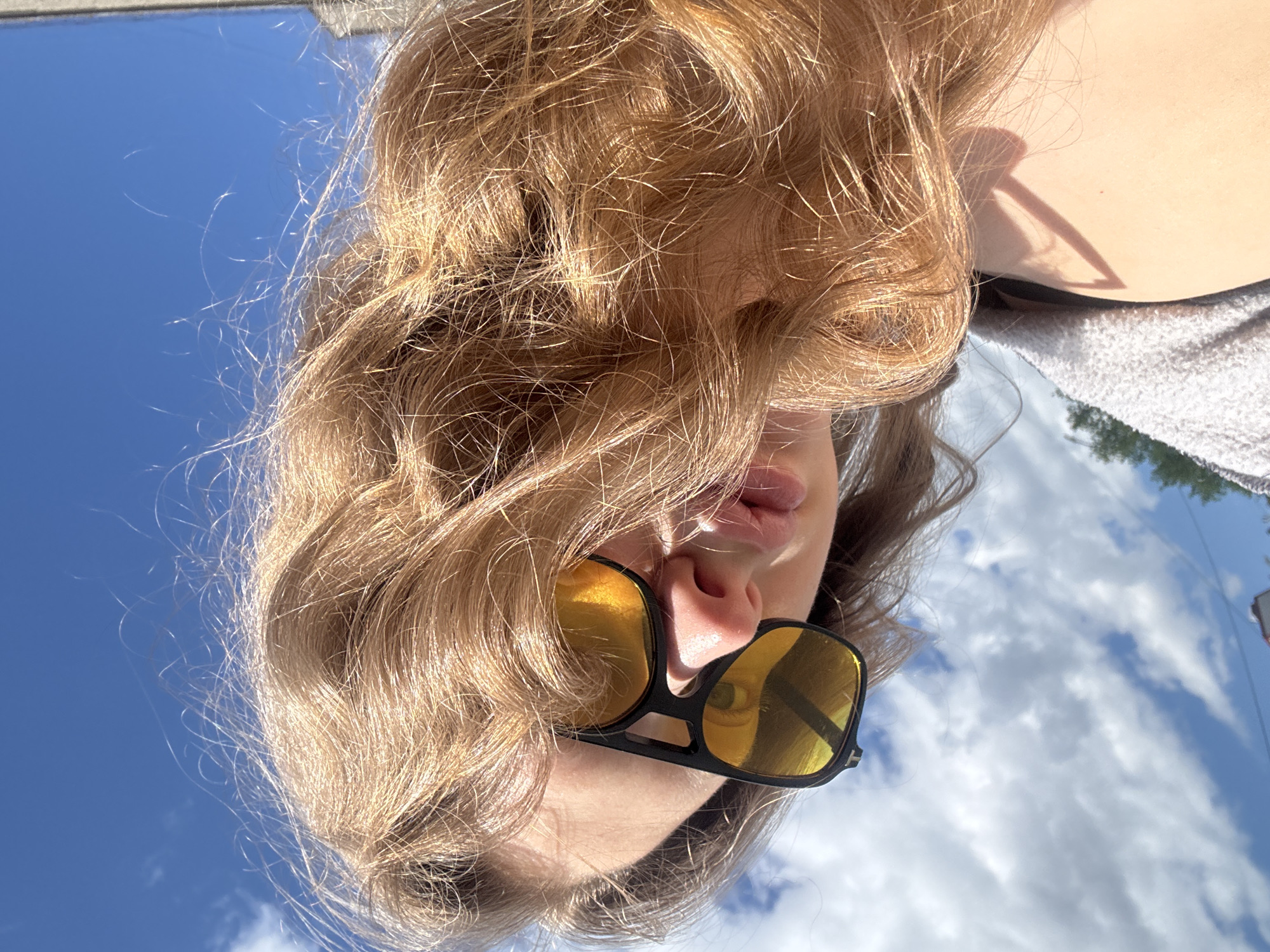 A young woman with blonde, wavy hair is looking up at the sky. She is wearing yellow-tinted sunglasses. The background is a bright blue sky with scattered white clouds. The image appears to be taken outdoors on a sunny day. There is no discernible location within the city of Izhevsk, Russia, as the background is solely sky and a hint of a building on the far right.