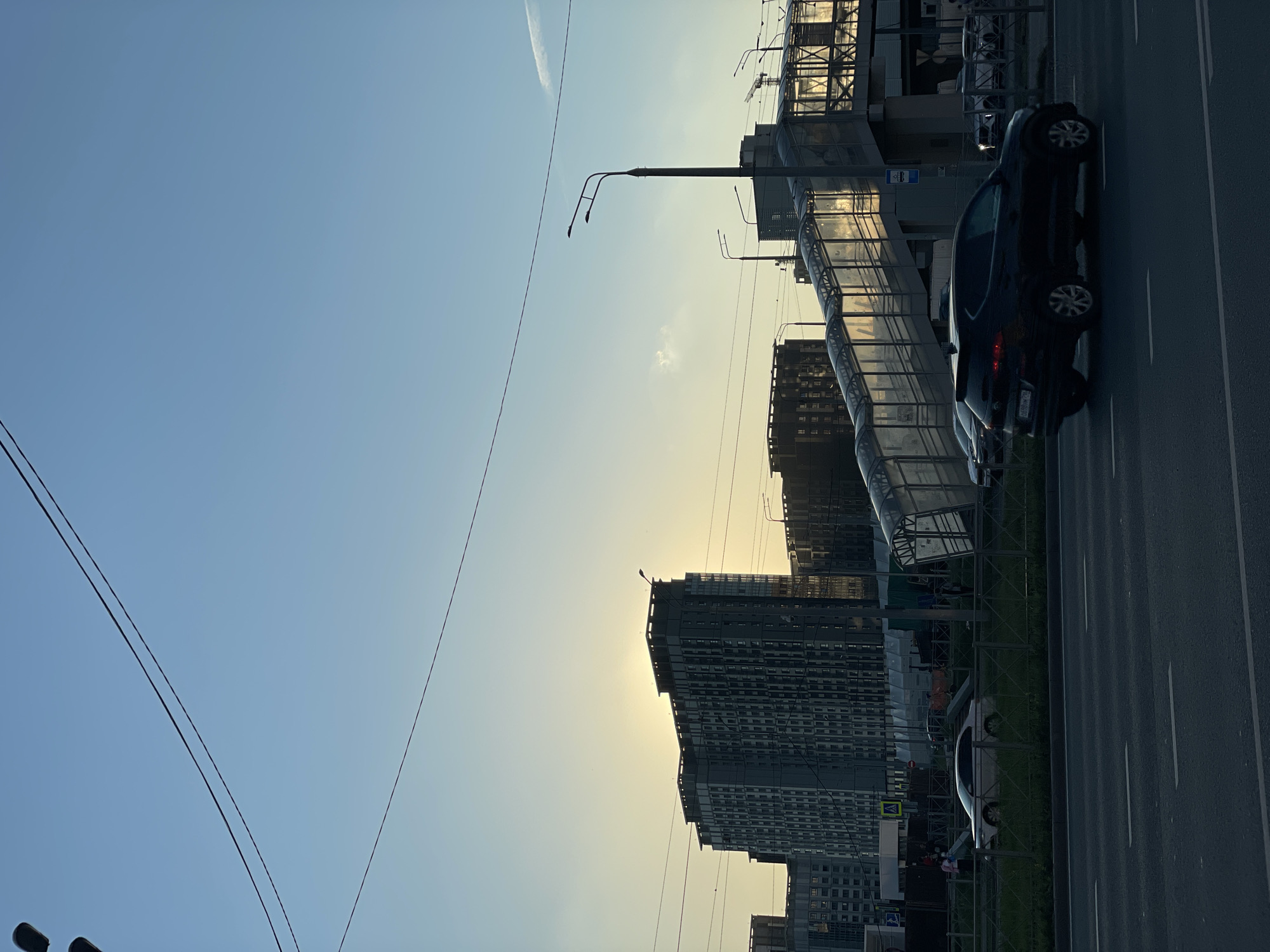 The image captures an urban street scene in Kazan, Russia, during either sunrise or sunset. The sky, transitioning from a light blue at the top to a pale yellow-orange near the horizon where the sun is low, dominates the upper left portion of the frame. A faint contrail and a small, wispy cloud are visible against the clear sky.
On the right side, a multi-lane asphalt road with white lane markings stretches across the foreground. A dark-colored sedan, possibly black or dark blue, is parked or moving slowly on the road. Nearby, a blue bus stop sign with a white bus icon is visible. Parallel to the road, a green metal fence separates the roadway from a grassy strip. Behind the fence, a lighter-colored car is partially visible.
Multiple tall, modern buildings, some appearing to be under construction or recently completed, rise against the horizon, their forms partially silhouetted by the low sun. The sunlight reflects off their facades and a prominent pedestrian overpass structure with transparent or mesh-like coverings. Two streetlights with multiple arms extend into the sky from the midground, and utility wires crisscross the upper part of the frame. The scene conveys a quiet moment in a contemporary urban landscape. No prominent human activity is discernible, and no legible text beyond the bus stop icon is visible.