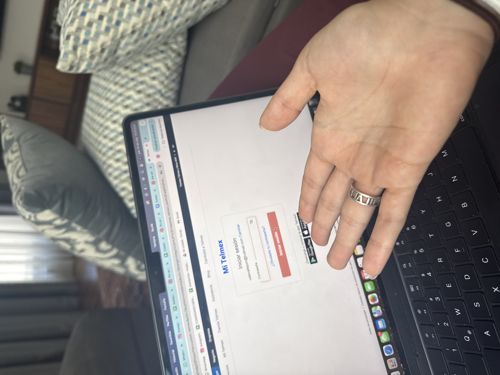 Here's a forensic description of the image:
A hand with a ring on the ring finger is positioned above a laptop keyboard. The laptop screen displays a login page for "Mi Telmex" with fields for username (valevallejom@hotmail.com) and password. The hand appears to be interacting with the screen. The keyboard layout is visible, and various browser tabs are open on the laptop. The background features pillows with a patterned design.
The location of San Bernardino Tlaxcalancingo, Mexico is not discernable from the image.