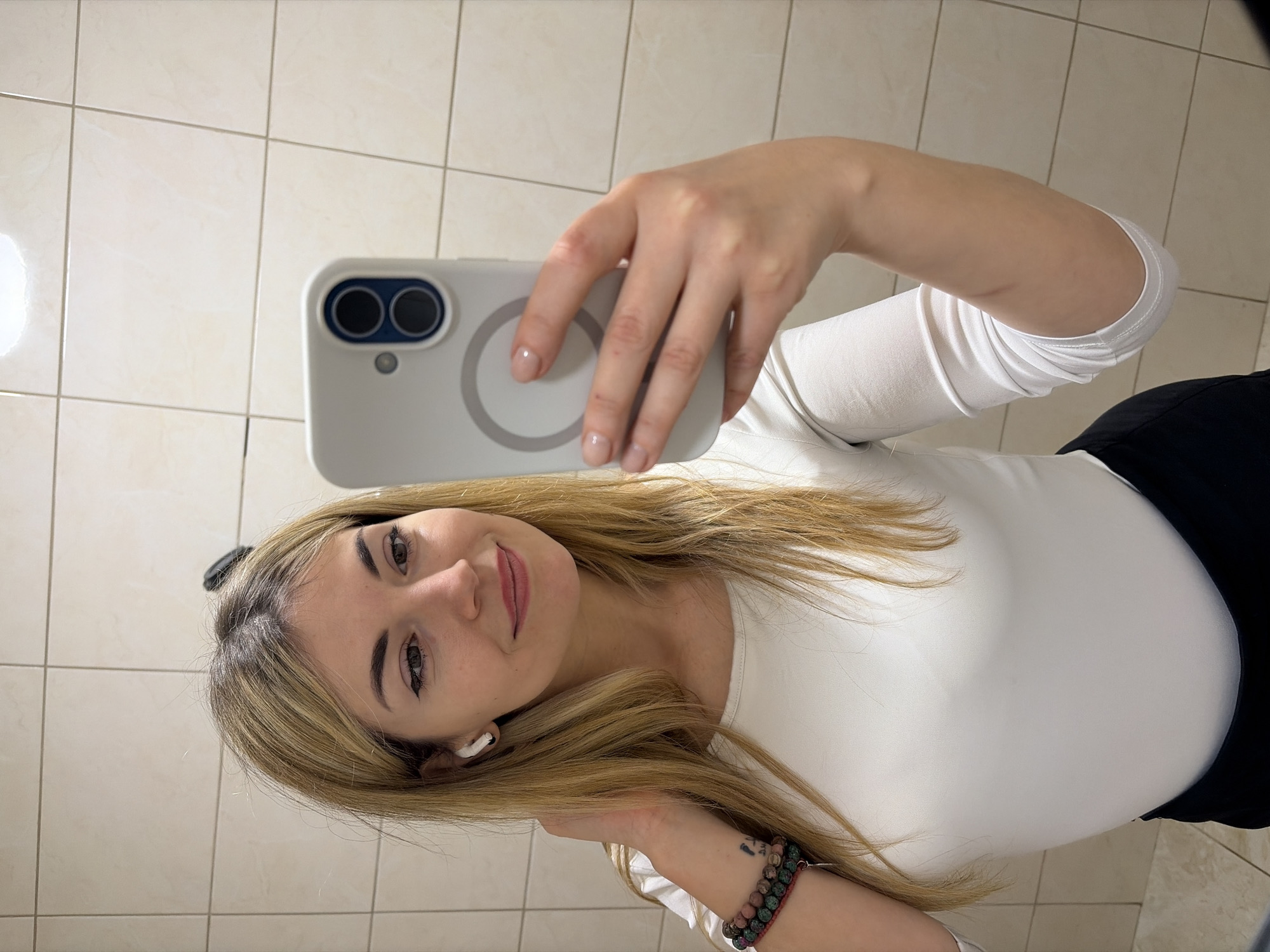 A woman with blonde hair and fair skin is taking a selfie in a bathroom. She is wearing a white long-sleeved shirt and black pants. She is holding a white smartphone with a circular magnetic attachment on the back. She is wearing an earbud in her left ear and bracelets on her left wrist. The background is a tiled wall. There is no evidence to support the location is the city of Budva, Montenegro.