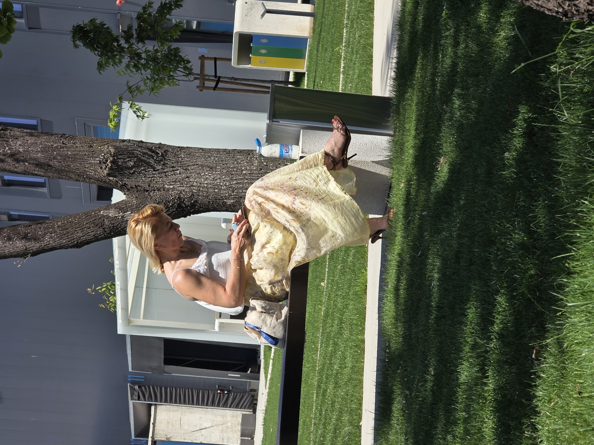 A person is seated on a bench outdoors, with their legs extended in front of them. The person appears to be using a mobile phone. They are wearing a light-colored dress and heeled shoes. Next to the person is a water bottle. The setting includes a tree, grassy areas, and in the background there are architectural elements such as a building and recycling bins. The picture was most likely taken in a park located in the city of Plovdiv, Bulgaria.