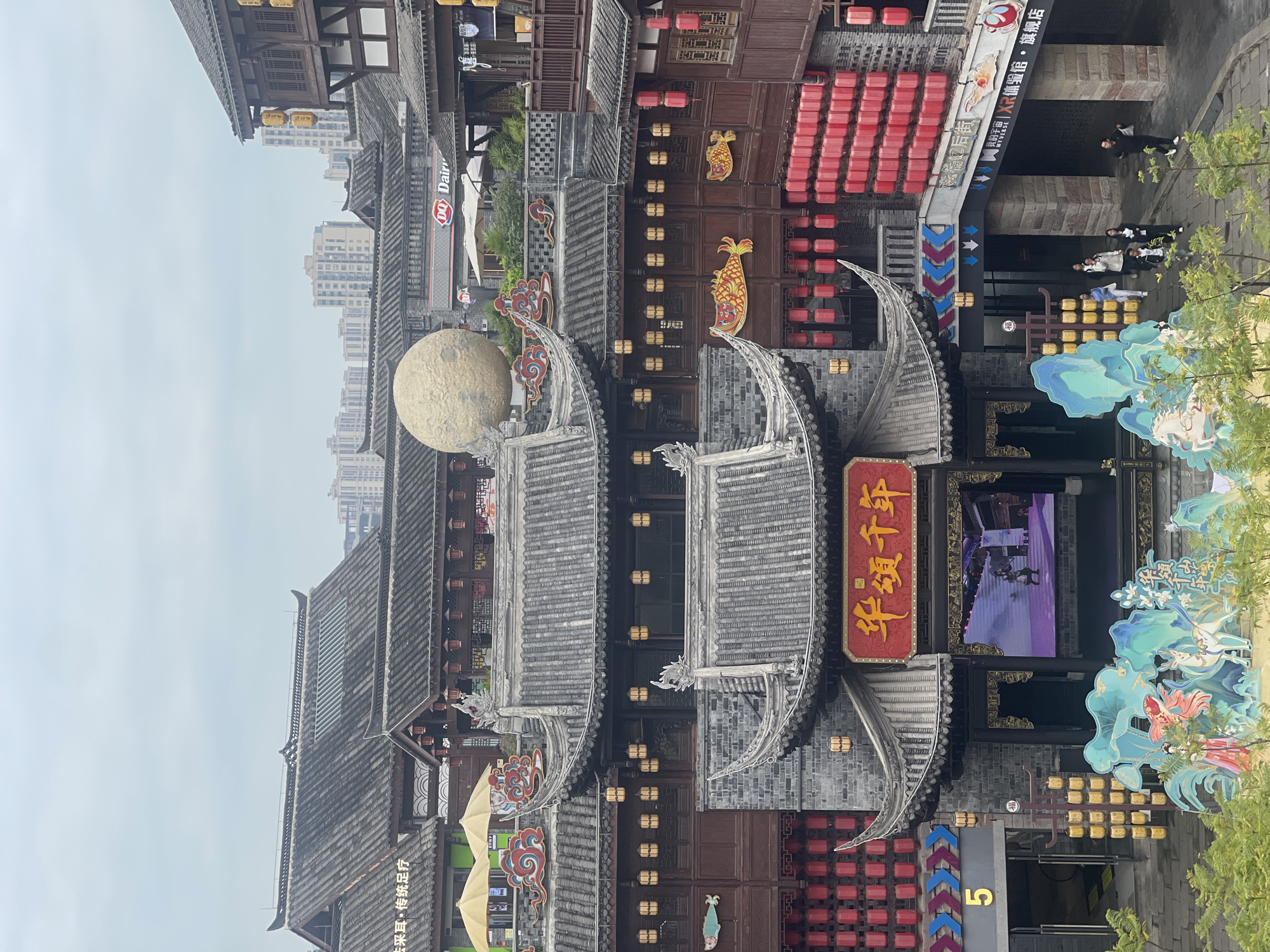 The image shows an urban scene with traditional Chinese architectural elements in Shapingba District, China. Buildings with gray tiled roofs and traditional curved eaves are prominent, adorned with red lanterns and decorative details. A large, spherical, moon-like structure is perched on one of the roofs. A modern DQ Dairy Queen sign is visible amongst the traditional buildings, blending the old with the new. The street is populated with a few people, mostly pedestrians. There's a shop with a red sign that reads "华颂千年" and the smaller text "感器口后街". Next to it, there is a sign of "XR体验馆·旗舰店", which means "XR Experience Museum - Flagship Store". The number "5" is visibly labeled on a building near a doorway. The signage "去采耳·传统足疗" is also visible, meaning "Ear Picking - Traditional Foot Massage". The environment is a mixture of old-style architecture and modern commercial elements.