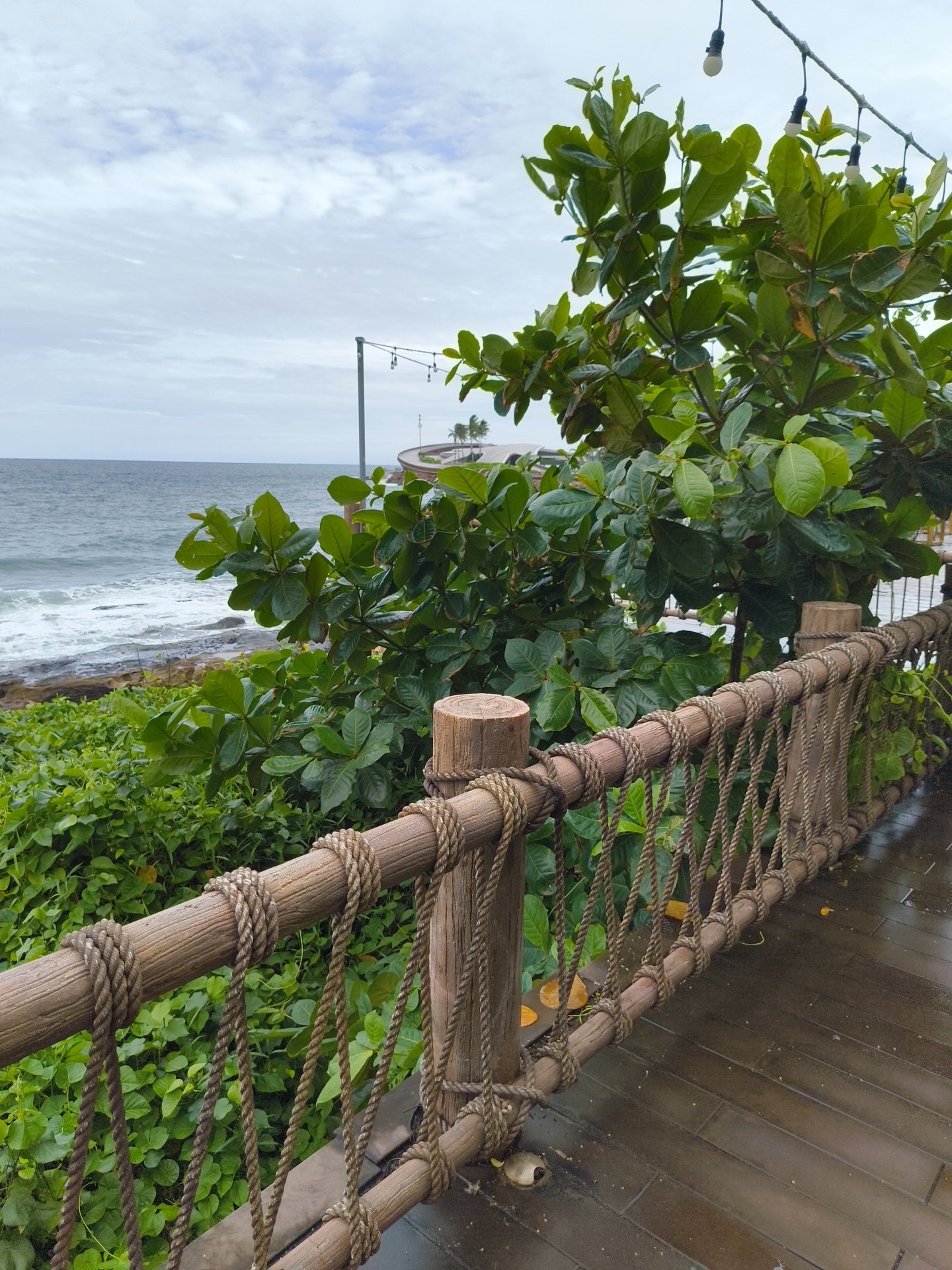 A weathered wooden railing with rope accents borders a wet wooden walkway. Beyond the railing, lush green foliage and a large tree with broad leaves dominate the foreground. In the background, the ocean meets a rocky shore under a cloudy sky. The scene appears to be a tranquil outdoor area, possibly a scenic viewpoint or walkway in Phú Quốc, Vietnam.