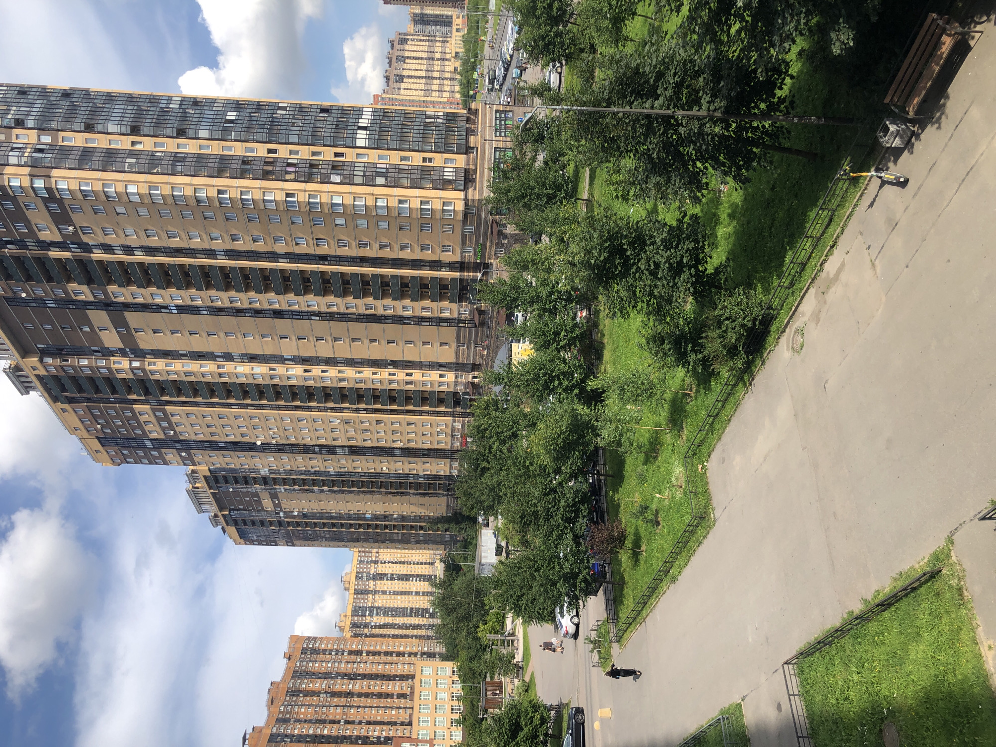 This is an outdoor, eye-level, medium-shot, daytime, vertical photograph taken in an urban environment with multiple high-rise apartment buildings dominating the background. The buildings are a mix of beige, light brown, and orange brick facades, with numerous windows and balconies. The sky is blue with scattered white clouds.
In the foreground and midground, there is a paved walkway and a grassy area with trees. A few cars are parked on the street visible in the distance. A person dressed in black is walking on the paved walkway. Another two people are walking on a different pathway in the midground, near a white car. Several other vehicles are parked and visible on the far right of the image.
The overall scene depicts a residential area within a city.