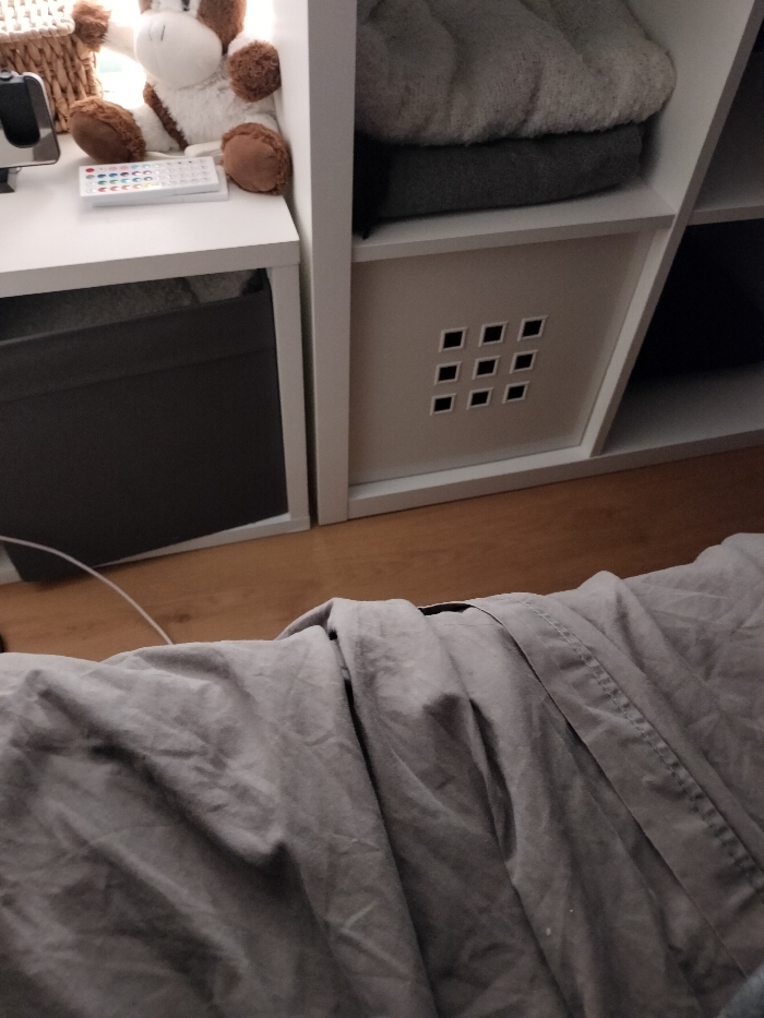 The image depicts an indoor scene in Cournon-d'Auvergne, France, observed from a low perspective, likely a bed. The lower half of the frame is dominated by rumpled, light grey fabric, presumably a duvet or blanket. Above this, a light brown wooden floor is visible, leading to two white modular cube shelving units.
On the left shelving unit, a white stuffed animal with brown patches (resembling a giraffe) is positioned on the top shelf, alongside a partially visible woven basket and a black electronic device. In front of the stuffed animal, a white remote control with multiple colored buttons rests horizontally. The lower compartment of this unit contains a dark grey fabric storage cube. A thin white cable extends from the lower left towards this area.
The adjacent right shelving unit contains two folded textiles in its upper compartment: a light grey fluffy item over a darker grey one. The middle compartment holds a light beige fabric storage cube featuring a nine-square grid cutout pattern. The bottom compartment of this unit is partially visible and dark. No individuals are present. The scene is illuminated by soft, ambient light.