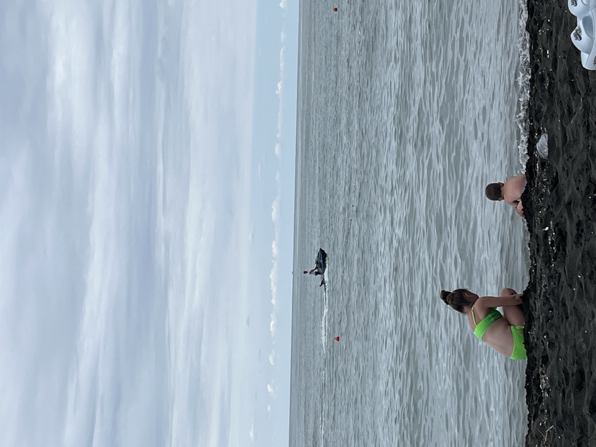 The image depicts a scene at Ureki, Georgia, a location known for its black magnetic sand beaches. The setting is a beach with dark sand meeting the calm, grey water of the sea under an overcast sky.
The subjects are primarily people enjoying the beach and water. In the foreground, a young girl in a bright green bikini is seated on the dark sand, with her legs drawn up to her chest. Further along the shore, a person in swim trunks is kneeling or crouching at the edge of the water. In the middle ground, a person is riding a jet ski, leaving a wake behind them on the water's surface. There are also a few red buoys visible in the water, likely marking a swimming area or course. In the upper right corner, a pair of white flip-flops are visible on the sand.
The time of day appears to be daytime, given the ambient light, but the cloudy sky suggests the sun is not directly visible. The weather is overcast, contributing to the muted tones of the water and sky. The overall mood is relaxed, with people engaging in typical beach activities. No text is visible in the image.