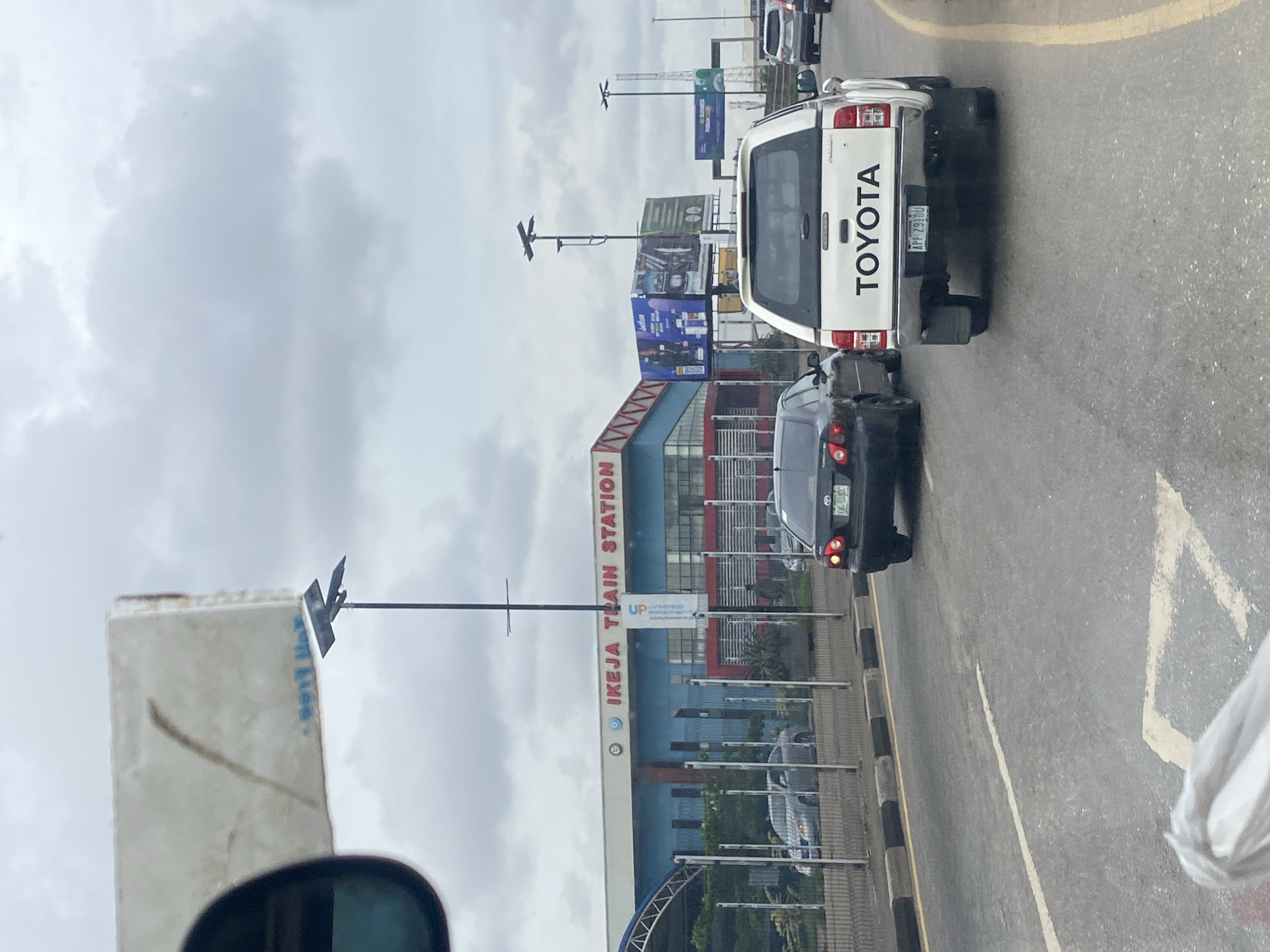 A medium shot captures traffic on a cloudy day in Ikeja, Nigeria, with the "IKEJA TRAIN STATION" sign prominently displayed on a blue and red building in the background. Several vehicles are visible on the asphalt road, including a white Toyota pickup truck in the foreground with a license plate reading "APP 2918U". A black sedan is in front of the Toyota, and another gray sedan is further ahead. Billboards are present on the right side of the frame. The environment appears to be an urban setting with infrastructure for transportation.