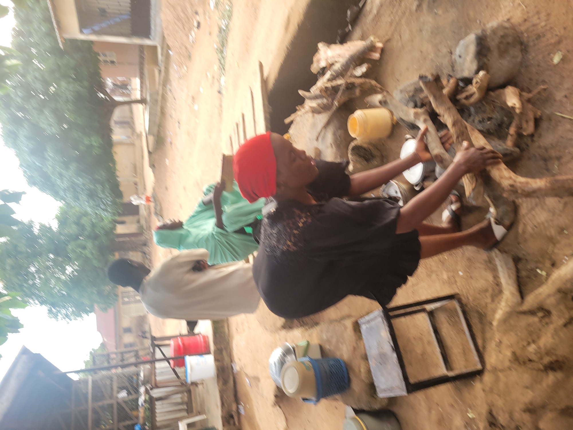 In Madalla, Nigeria, a scene unfolds outdoors with a woman wearing a red headscarf and black clothing bent over a makeshift stove. She is tending to a fire fueled by wood, with her hands positioned near the burning logs. To her right, a processed animal carcass, possibly a goat, lies on the ground near the fire. In the background, other individuals are present. A man in a light-colored garment stands facing away from the camera, while another person in a green veil sits. Various household items, including pots, buckets, and a yellow container, are scattered around the immediate area. The ground is composed of dry earth and sand, indicative of a dusty environment. Buildings and trees are visible in the distance, suggesting a residential or market setting within the town.