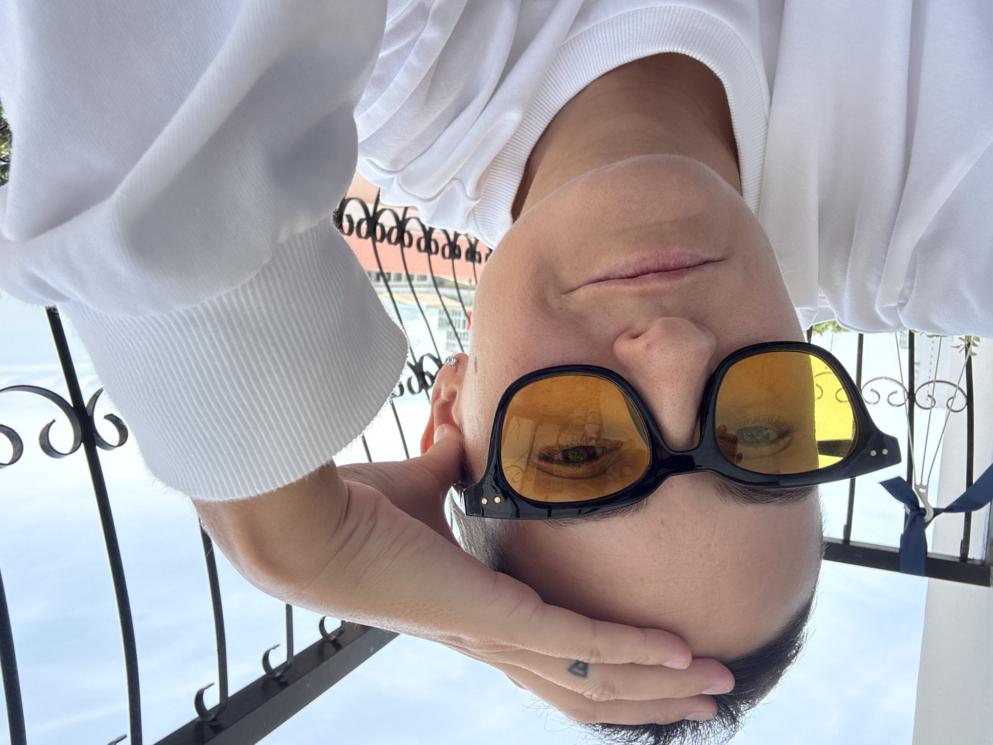 A person with short hair and sunglasses with yellow lenses is taking a selfie on a balcony in Nha Trang, Vietnam. The person is wearing a white sweatshirt and has their hand on their forehead. The background shows a railing and a blue sky.