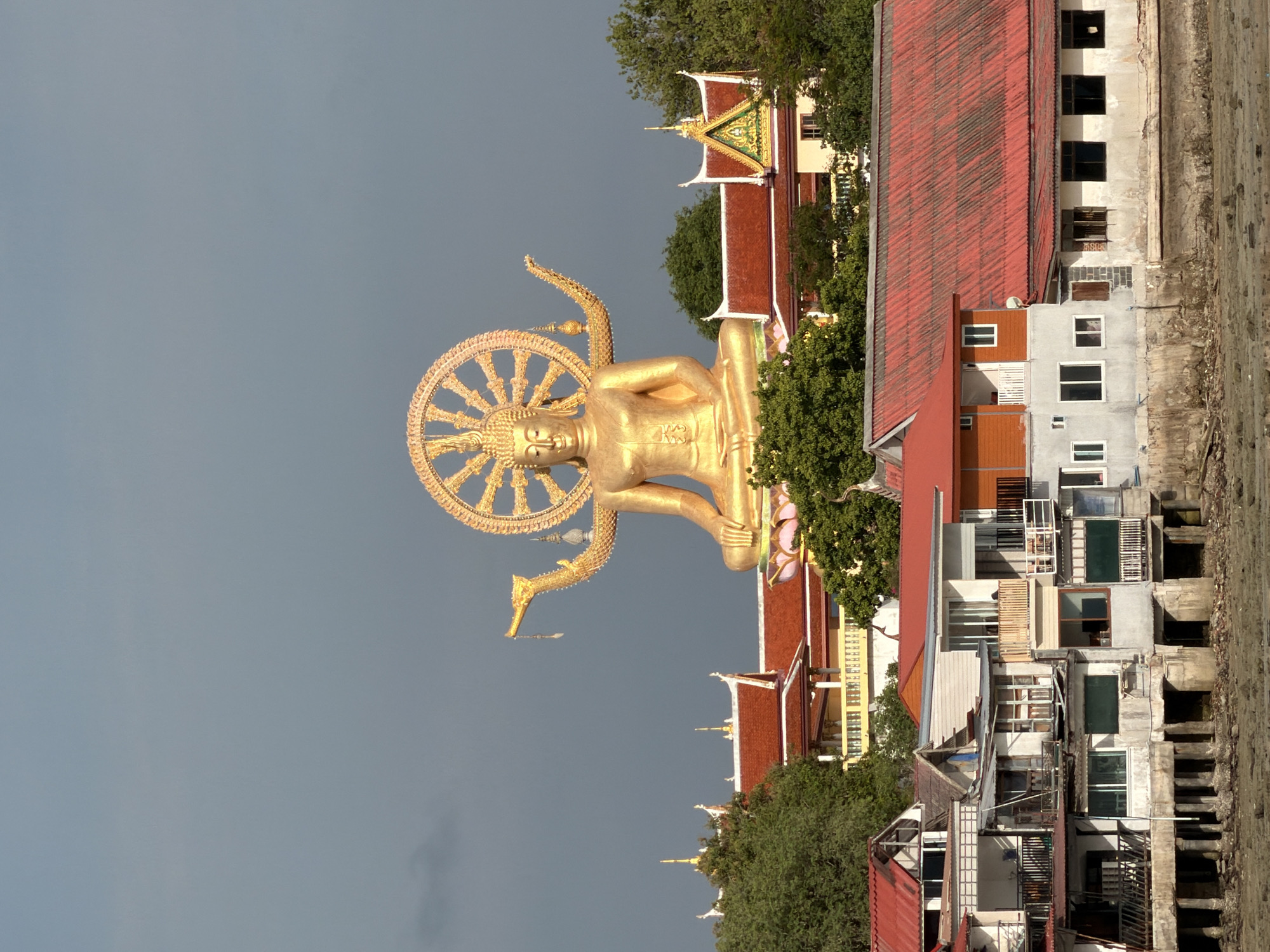 The image shows a large golden Buddha statue overlooking a waterfront village on Ko Samui, Thailand. The statue is seated with its legs crossed and has a halo behind its head, which is adorned with a circular motif. The Buddha has multiple arms, each holding or gesturing in a specific way. Behind the statue, several buildings with red tiled roofs and ornate architectural details are visible, suggesting a religious or temple complex. In the foreground, a cluster of buildings with varying architectural styles, including some that appear to be overwater structures with visible pilings, are lined up along the water's edge. The water appears shallow and muddy, with some rocks exposed. The sky is overcast and gray, creating a somewhat dramatic backdrop for the scene. The overall impression is one of a juxtaposition between a prominent religious landmark and a more humble, everyday residential and commercial area.