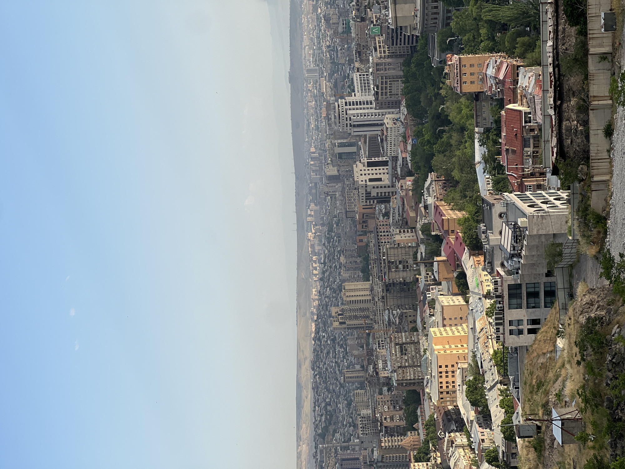 The image is a high-angle, wide shot of a sprawling cityscape in Yerevan, Armenia, under a clear blue sky with a hazy horizon. The foreground features a dry, grassy hillside with rocks and a small, weathered structure. The middle ground and background are densely populated with buildings of varying architectural styles and heights, ranging from older, multi-story residential blocks to more modern, concrete structures and some with reddish-brown roofs. Green trees are interspersed throughout the urban landscape, particularly in the mid-ground. The distant background shows a range of hills and mountains, fading into a hazy atmosphere. There are no people or animals visible in the image. The overall impression is of a vast urban area in a somewhat arid environment, likely taken from an elevated viewpoint within the city.