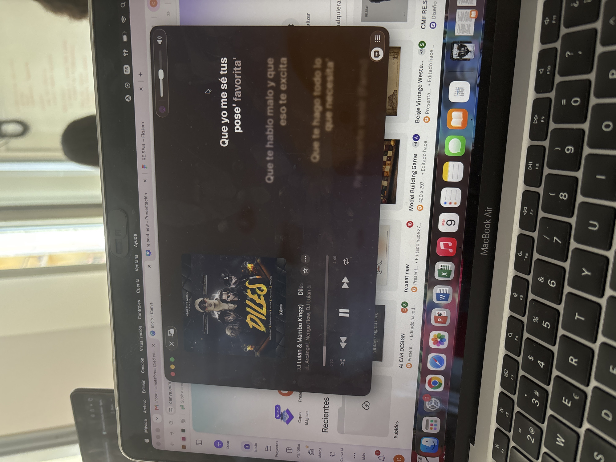 A close-up, overhead shot shows a MacBook Air laptop with a music player open on its screen. The music player displays album art with the title "DILES" and lyrics in Spanish. The laptop's keyboard and a dock of application icons are visible. The background is out of focus, showing a blurred interior environment with hints of windows and a possibly urban setting. There is no indication of Milan, Italy, or any specific city.