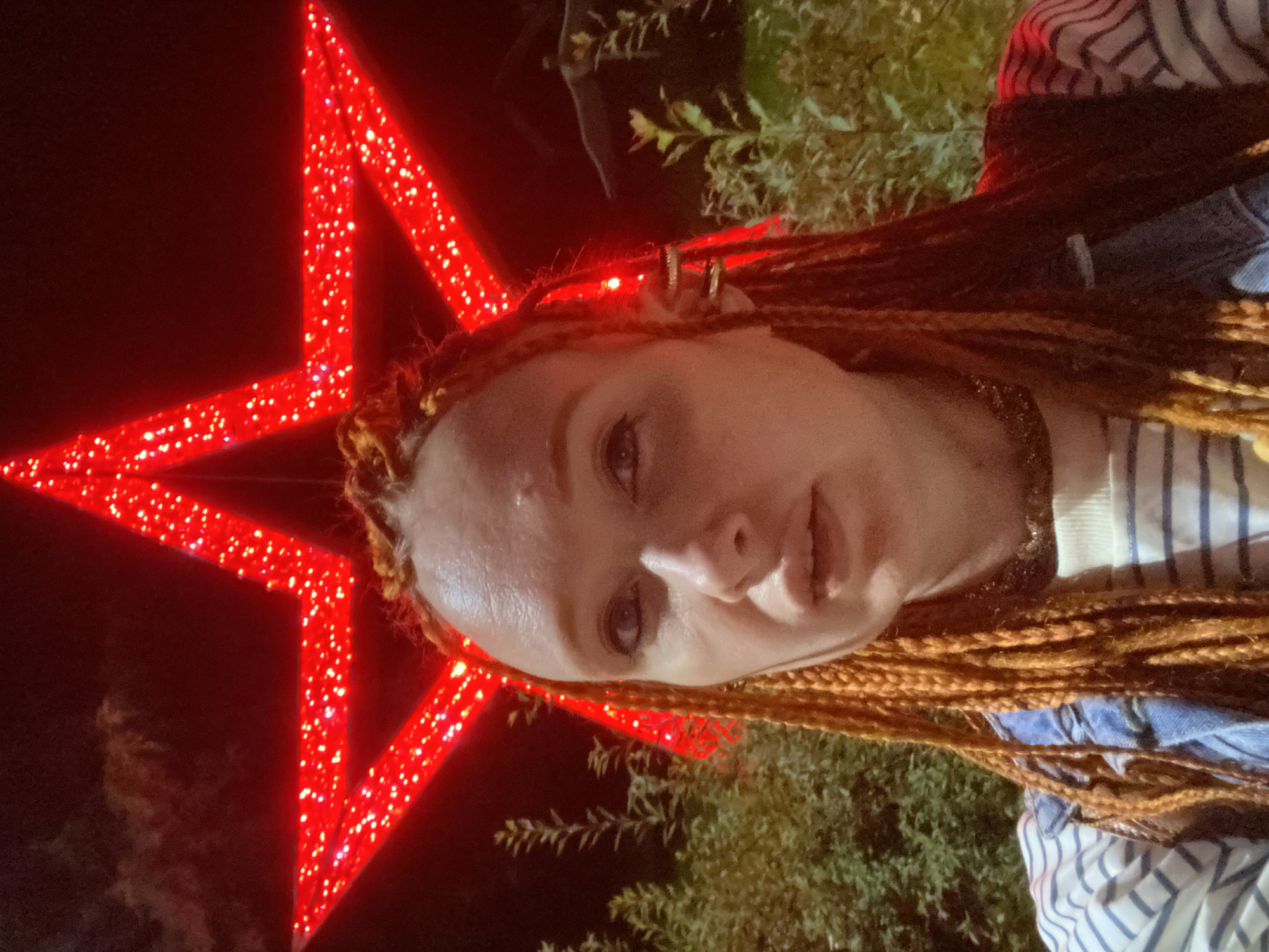 An individual with auburn braided hair is captured in a selfie outdoors at night, with a large, illuminated red star decoration prominently positioned behind them. The star is made of red lights embedded in a frame, creating a shimmering effect. The background includes dark foliage and what appears to be a building structure. The individual is wearing a striped shirt and a denim jacket. Their face is turned slightly towards the camera, and their expression is neutral. The scene suggests a festive or decorative outdoor setting, potentially in the city of Sochi, Russia, given the context of the query.