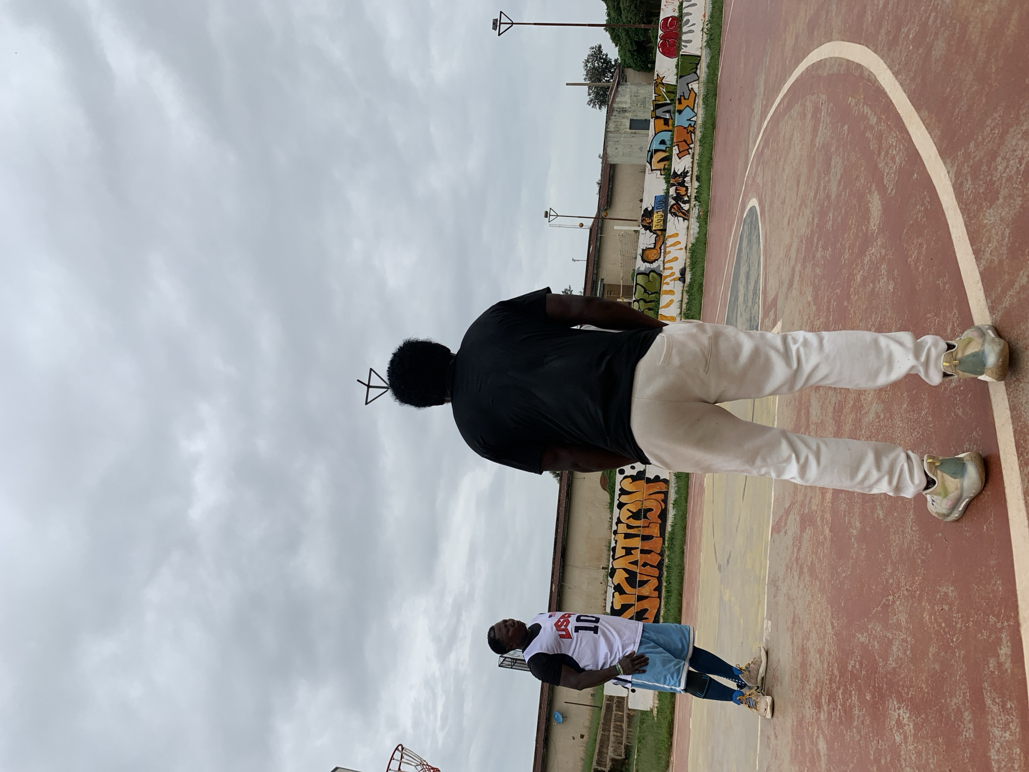 Two men are on a basketball court under an overcast sky. One man, on the left, wears a white basketball jersey with red trim and the number "10" in blue, and light blue basketball shorts. He stands facing the other man, looking to his right. The second man, on the right, faces away from the camera, wearing a black t-shirt and light-colored sweatpants. He is positioned centrally in the frame. The basketball court has a reddish-brown surface with painted white lines. Behind the players is a low wall covered in graffiti. In the background, there are some buildings and trees. The overall atmosphere is that of an outdoor recreational area, possibly a park or a school yard. The scene appears to be located in Jos, Nigeria, given the context provided.