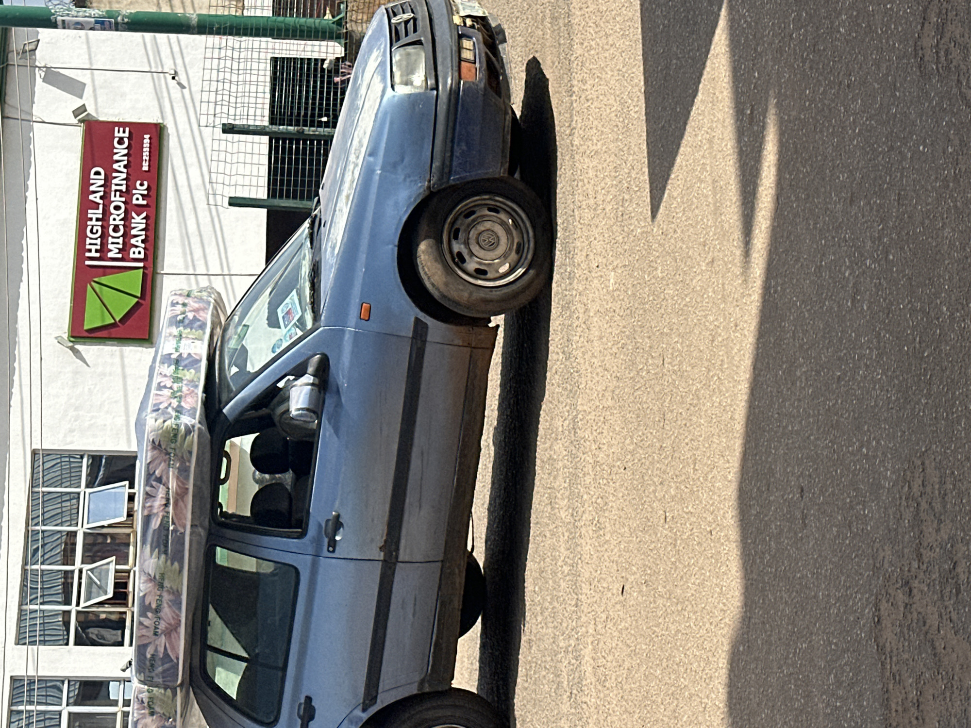 In Bukuru, Nigeria, a light blue Volkswagen sedan is parked on a paved street. The car is transporting a large mattress secured to its roof. The mattress is wrapped in clear plastic with a floral pattern. In the background, a building with large windows displays a sign for "HIGHLAND MICROFINANCE BANK Plc." The environment appears to be a commercial area with a paved road and a sidewalk. The scene is captured during daylight hours, with shadows cast on the street.