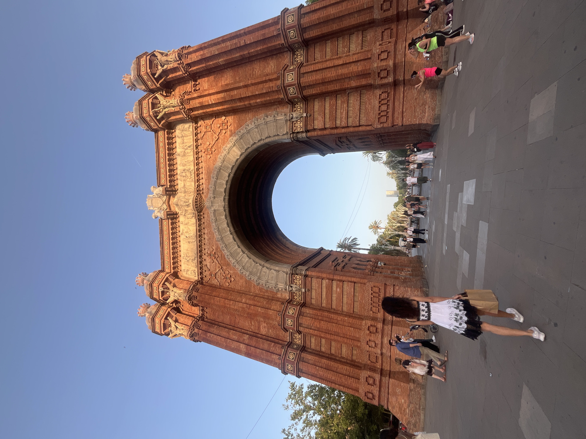 The image shows the Arc de Triomf in Barcelona, Spain. It is a triumphal arch made of brick. In the foreground, a woman in a white dress with black trim is walking away from the camera, carrying a brown paper bag. Several other people are visible in the background, walking or standing near the arch. The sky is clear and blue, and palm trees are visible behind the arch. The scene appears to be in a public square or park, with paved ground and a few trees.