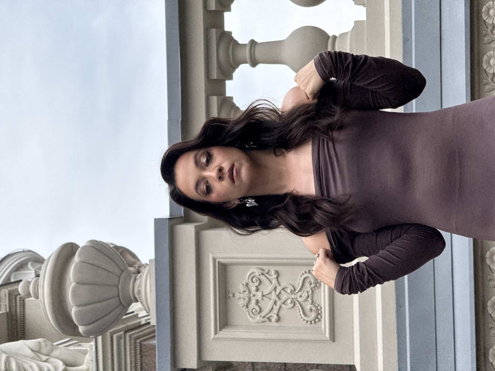 A medium shot captures a woman in an off-the-shoulder, dark brown, long-sleeved dress, posing outdoors against a backdrop of architectural elements. She stands on what appears to be a balcony or terrace, with ornate balustrades and decorative stonework behind her. The sky above is overcast. The woman's dark, wavy hair falls past her shoulders, framing her face. She wears eye makeup with a winged liner and light-colored eyeshadow, and her lips are slightly parted. A heart-shaped earring dangles from her left ear, and she wears at least two rings on her right hand, which is casually placed on her shoulder, adjusting the sleeve of her dress. The architectural details suggest a historical or grand building, potentially in an urban setting. The location is identified as Kazan, Russia.