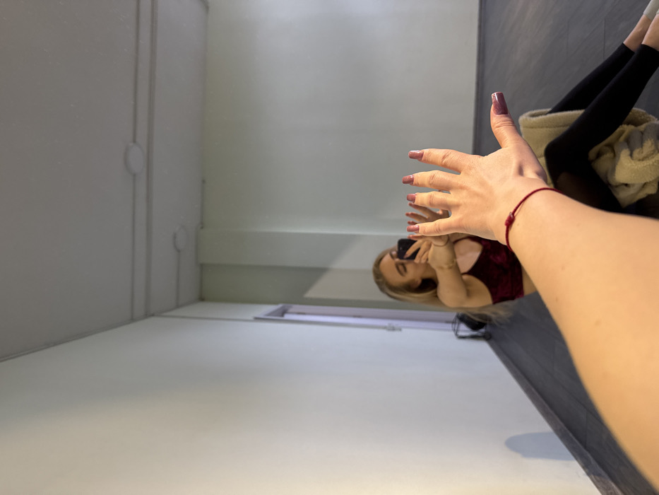 The image portrays a mirror selfie taken inside a studio or multi-purpose room in the city of Unknown, Unknown. A blonde-haired individual, presumed female, is seated on a dark gray floor, facing a large wall-mounted mirror. She wears a dark red, velvet-textured crop top and dark leggings, with a light-colored towel draped across her lap. The individual is holding a black smartphone with both hands, aiming it at the mirror to capture her reflection. In the immediate foreground, the photographer's extended left arm is prominent, positioned towards the mirror with fingers slightly splayed. A red string bracelet is visible on the wrist of this arm, and the fingernails are painted with a muted pink or mauve polish. The interior environment, visible in the reflection, features light pale green walls, a light-colored doorframe with a dark interior on the left, and a white ceiling equipped with circular light fixtures and linear conduits.