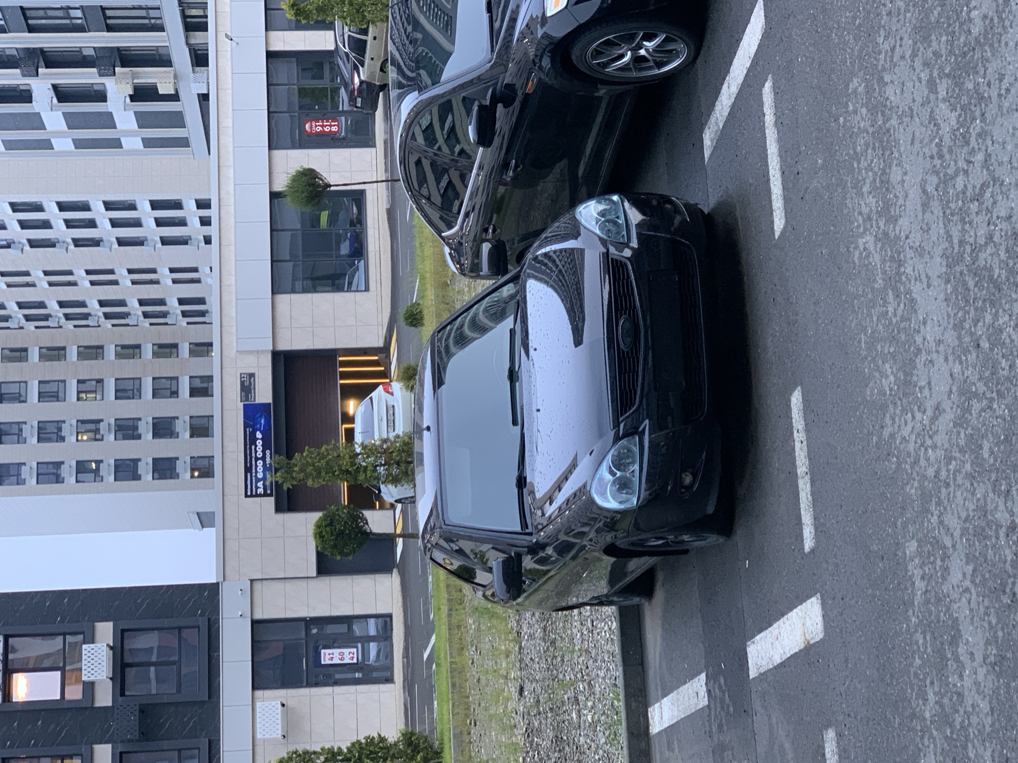 Two black cars are parked in a lot in front of a modern building. The car in the foreground is a dark-colored sedan, possibly a Lada Granta, facing slightly to the right. It has water droplets on its surface, indicating recent rain. The second car, a black Ford Mondeo, is parked to the right of the first car, also facing right. Both vehicles are in designated parking spots marked with white lines. The building behind the cars features large windows and balconies. A white sedan is visible driving out of an underground parking entrance in the background. The environment appears to be a city street or residential area. The building's signage includes Russian text, suggesting the location is in Russia. Based on the Cyrillic text "3A 600 000 Р" on a banner above the parking entrance, which translates to "For 600,000 RUB", and other visible Cyrillic text, this scene is likely in Stavropol, Russia.