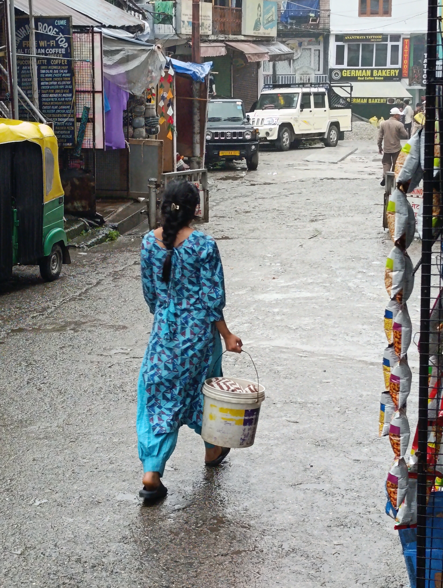 The image depicts a street scene in Vashisht, India, likely during monsoon season given the wet asphalt and overcast sky. In the foreground, a young woman walks away from the camera, carrying a white bucket in her right hand. She is wearing a blue and white patterned kurta and blue pants, and her hair is braided and tied back with a gold clip. The bucket contains items with a brown and white striped pattern. To the left of the woman is a green and yellow autorickshaw. In the background, shops line the street, with awnings and signage visible. One shop is identified as "ROOFTOP COFFEE" and another as "GERMAN BAKERY". A white SUV is parked further down the street. A man in a light brown shirt and dark pants is walking towards the bakery. The overall impression is a bustling, albeit wet, urban environment.