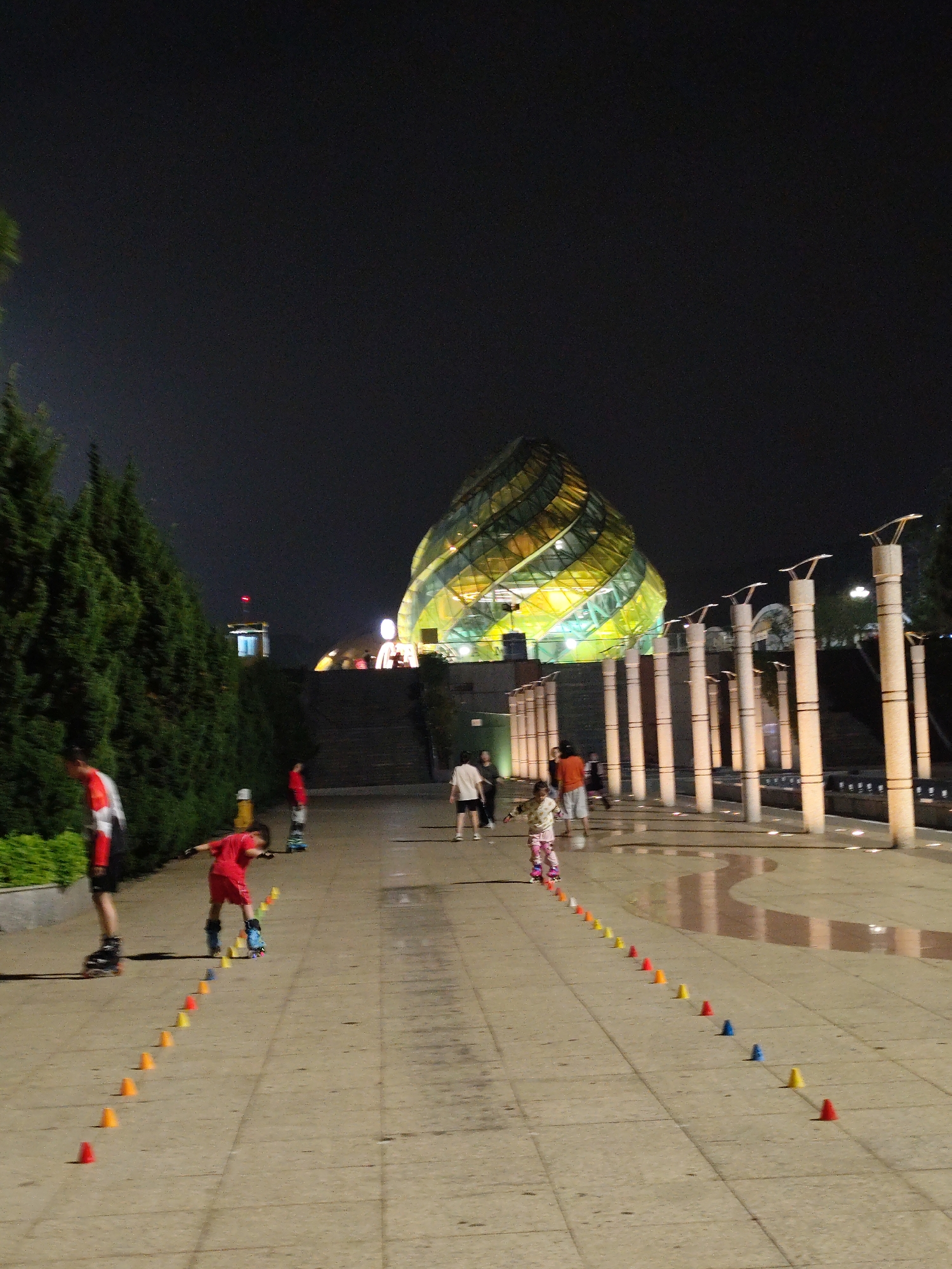 The image depicts a night scene in Da Lat, Vietnam, with people rollerblading along a walkway. In the background, there is a large, illuminated, geometrically-shaped building, likely the Da Lat Market's iconic "Artichoke" building. A line of small cones is arranged along the walkway to guide the rollerbladers. Several people of varying ages are visible in the scene, some wearing roller skates and others walking. Vertical columns with illuminated tops are present on the right side of the walkway. A row of bushes lines the walkway to the left.