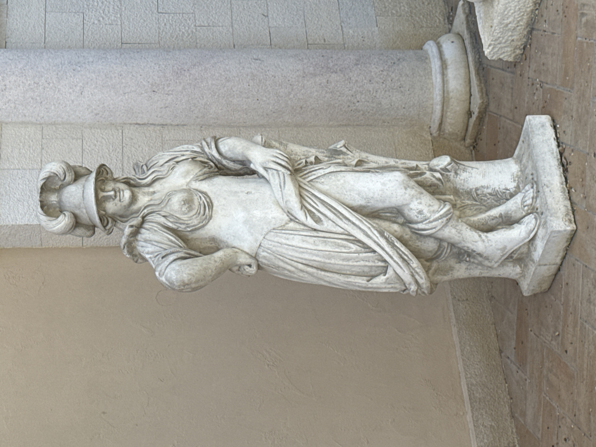 The image features a classical-style stone statue of a female figure, depicted in a reclining or semi-reclining pose. The statue is crafted from a light-colored material, likely marble or limestone, exhibiting fine detail and some signs of age or weathering.
The central subject is the female figure, portrayed with a serene expression. She wears an elaborate helmet or headpiece adorned with prominent, decorative curls or volutes. Intricately carved, wavy hair flows from beneath her helmet, framing her face and cascading over her shoulders. Her body is draped in flowing garments that gather in realistic folds, revealing parts of her chest and limbs. One arm is bent and rests across her torso, while the other is also bent at the elbow. Her bare feet are visible, protruding from beneath the drapery. The statue rests on a simple, rectangular base.
The setting appears to be an architectural space. To the left of the statue, a plain, light beige wall with a slightly textured finish forms the background. To the right, a substantial, rough-hewn stone pillar or column stands, suggesting a structural element of a building or arcade. Below and to the far right, a section of a tiled floor, in warm reddish-brown tones, is visible. The lighting is soft and diffuse, indicating either an indoor setting with natural light or an overcast outdoor environment, and casts gentle shadows that enhance the statue's sculptural depth. There is no discernible activity taking place, as the image captures a static artistic display. No text is visible in the frame. The overall impression is one of timeless art within a structured, possibly historical, environment in Varna, Bulgaria.