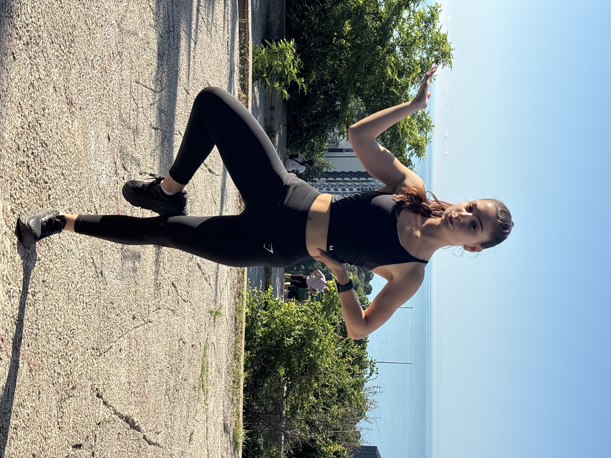 A young woman in black athletic wear, including a sports bra and leggings, is performing a yoga pose on a concrete surface with a cracked texture. She is balanced on her right leg, with her left leg bent and the sole of her foot resting against her right calf. Her left arm is extended upwards and outward, palm facing the sky, as if presenting something. Her right hand is on her hip. Her hair is pulled back in a ponytail.
In the background, lush green trees and bushes line the foreground. Beyond the greenery, a body of water, likely the Black Sea, stretches to the horizon under a clear blue sky. In the middle ground, between the trees and the water, faint outlines of buildings are visible, suggesting an urban environment. There are also a couple of other individuals in the background, further away from the main subject. The lighting indicates it is daytime, with a clear, sunny sky. The scene appears to be an outdoor park or promenade overlooking the sea. This location is consistent with areas along the coastline of Varna, Bulgaria, such as the Sea Garden or other elevated park areas offering views of the Black Sea.
