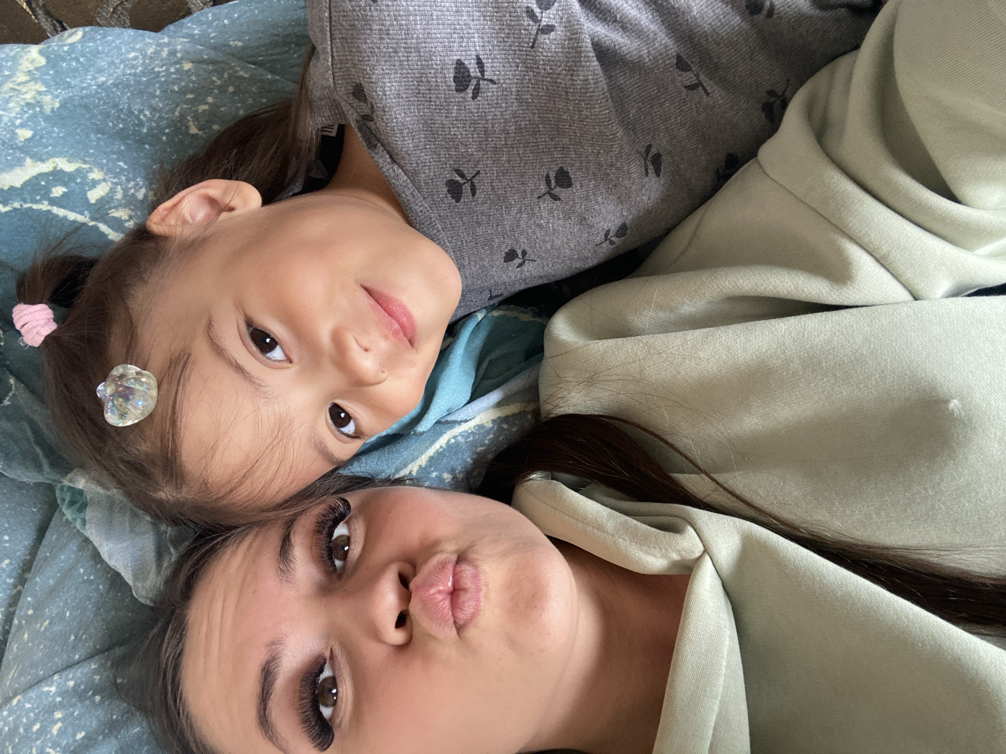 A photograph shows two individuals, one child and one adult, lying on a bed. The child is positioned above the adult, both oriented towards the camera. The child is wearing a gray shirt with a floral pattern and has a hair clip in their hair. The adult, presumed to be female, is wearing a light green sweater. The setting appears to be an indoor environment, on a bed with a blue patterned blanket. There is no information available to establish the location as Zhigalki, Russia.