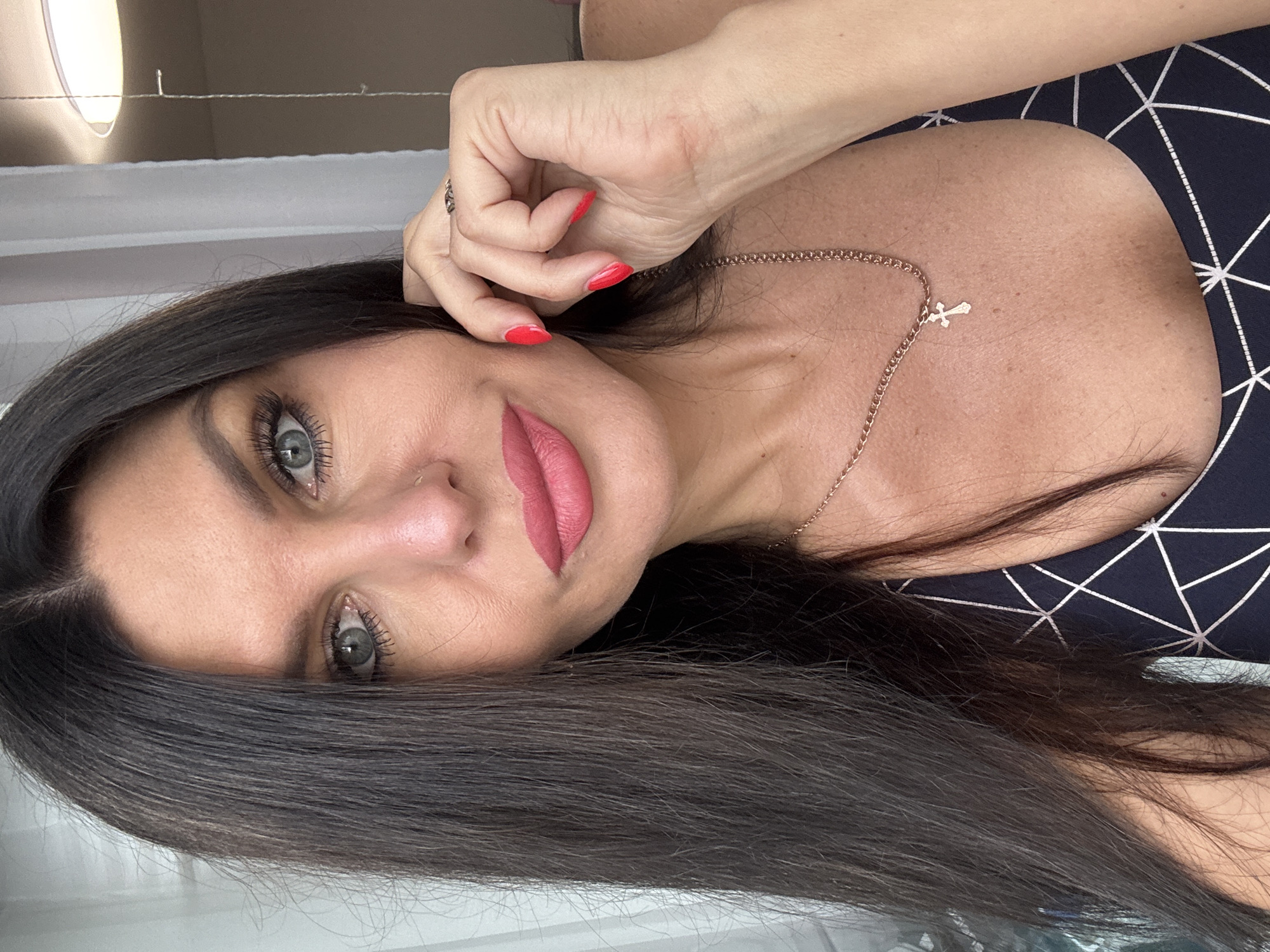 A close-up, waist-up shot shows a fair-skinned woman with dark brown hair, styled long and straight. She has bright blue eyes and is wearing a dark blue and white patterned top. Her lips are painted a matte pink, and her nails are a vibrant red. A thin gold chain with a small cross pendant rests on her chest. Her right hand is raised to her cheek, with a ring visible on her ring finger. The background is blurred, with sheer white curtains and a ceiling light suggesting an indoor setting. The image does not provide any information about a specific location in Chaikovsky, Russia.