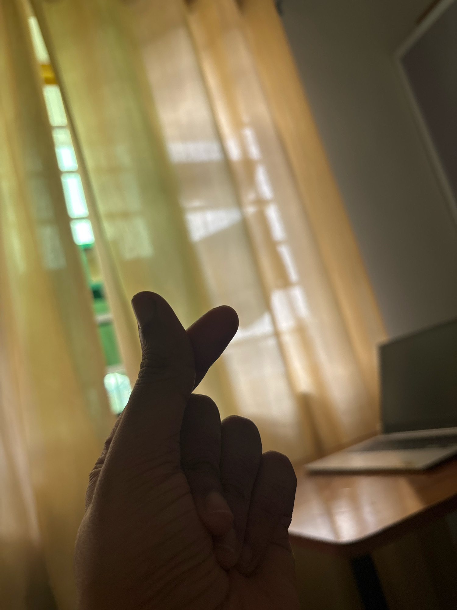 In Khalagon, India, a close-up shot focuses on a person's hand in the foreground, forming a heart shape with their thumb and index finger. The hand is positioned against a backdrop of sheer, light yellow curtains, through which sunlight streams, creating vertical light patterns. To the right, a wooden table is visible, with a blurred image of a laptop resting on it. The overall lighting is soft, with shadows indicating a light source from the left.