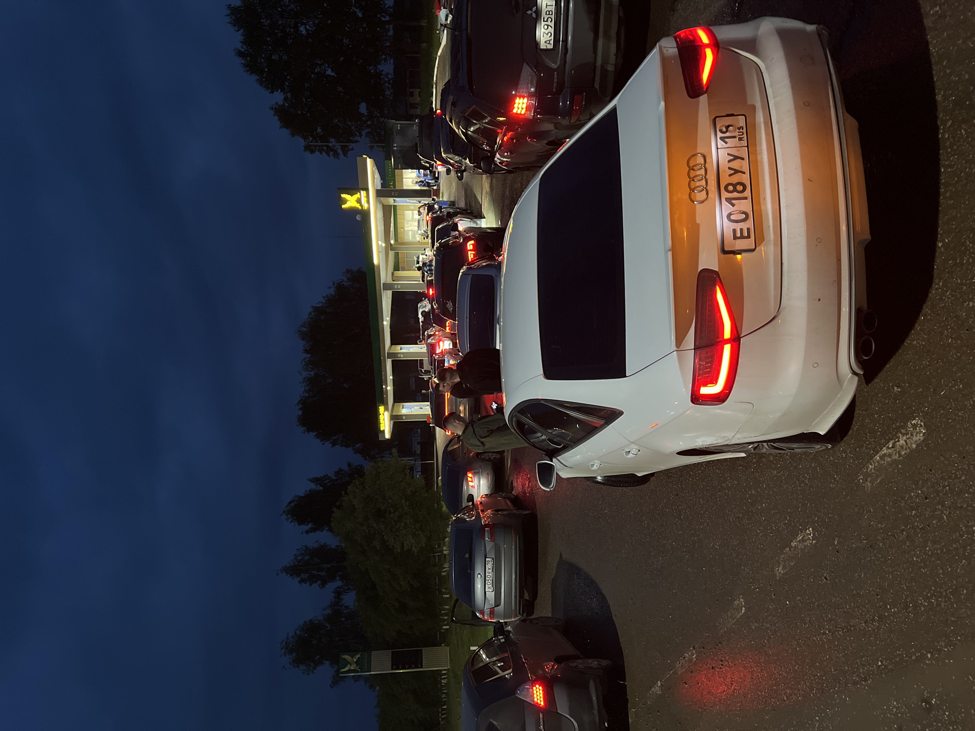 The image depicts a line of cars at a gas station in Agryz, Russia, during dusk or early evening. The sky is a deep blue, suggesting twilight or a slightly overcast evening. The gas station, illuminated by bright lights, has a prominent yellow "X" logo. Several cars are lined up, indicating a wait, possibly for fuel.
In the foreground, a white Audi with a partially visible license plate "E018yy 18 RUS" is parked. Further down the line, other vehicles are visible, including a dark SUV with a license plate starting with "FA395BT". People can be seen inside and around some of the cars, though their faces are not clearly discernible. The overall atmosphere appears ordinary, a typical scene at a busy gas station. The road surface is wet, hinting at recent rain or damp conditions. The presence of trees in the background adds to the outdoor setting. There is a sign with a bird-like emblem near the left side of the image.