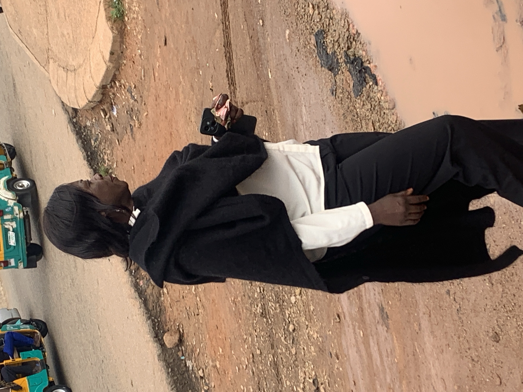The image shows a woman standing on a dirt road with a puddle of muddy water to her right. She is wearing a white shirt, black pants, and a black shawl draped over her shoulders. She is holding a smartphone and some money in her right hand. Behind her, two tuk-tuks are visible on the road, one of which is yellow and green and the other is green and white. The ground is a mixture of dirt, gravel, and some sparse vegetation. The scene appears to be in Gura Top, Nigeria, given the context of the vehicles and the environment.