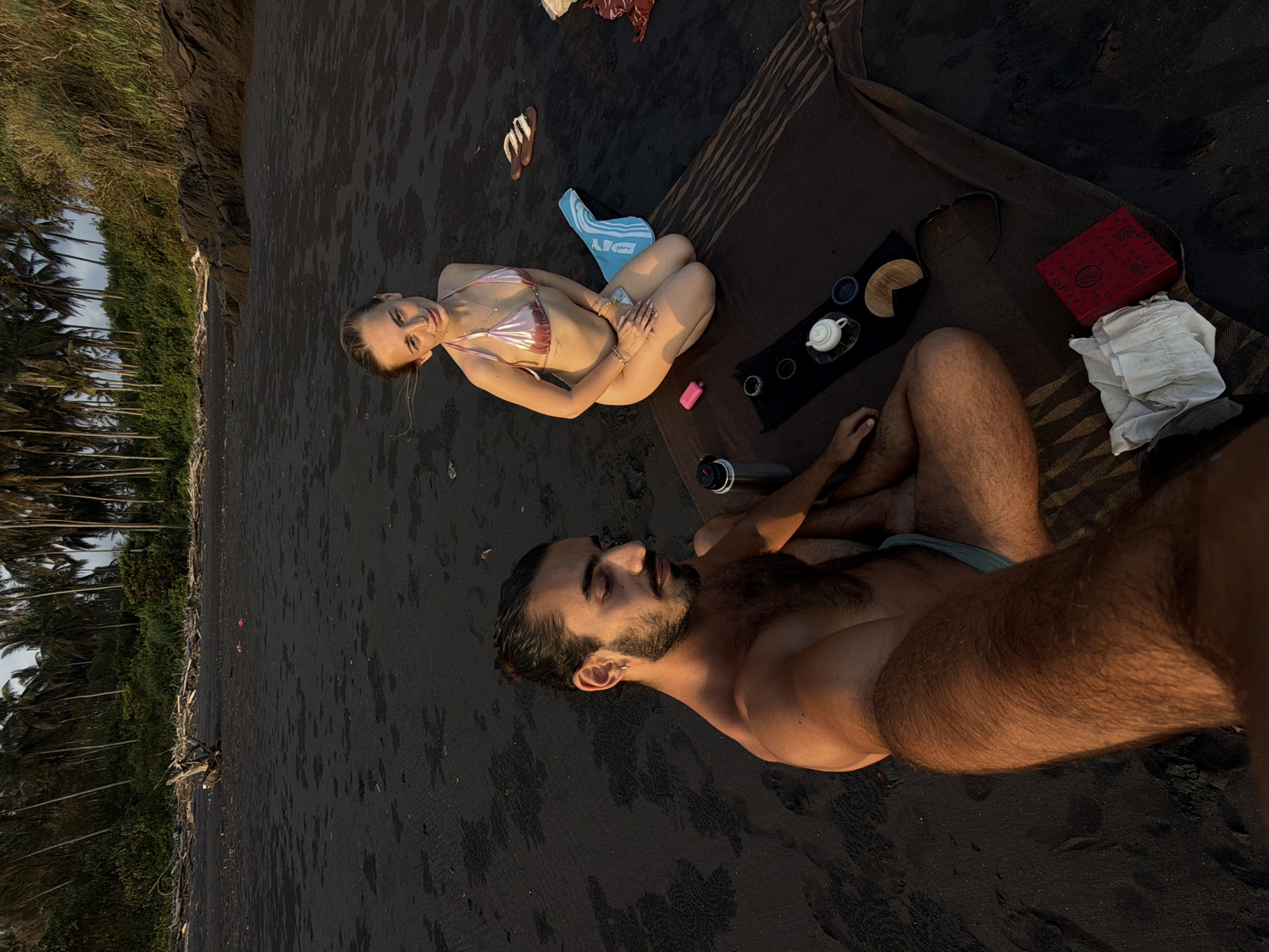 The media file captures a leisurely scene on a black sand beach in Selemadeg Timur, Indonesia, likely during late afternoon or early evening, indicated by the warm, low-angled sunlight casting long shadows and illuminating the subjects.
Two individuals, a man and a woman, are relaxing on a striped blanket laid out on the dark sand. The woman is positioned on her side, wearing a shimmering pink bikini. Her body is angled towards the camera, and her head is turned to look directly at the viewer with a relaxed, perhaps playful, expression. She is wearing bracelets on her wrists. To her side, a pair of brown flip-flops and a blue towel with white stripes are visible. Near her, a pink e-cigarette is placed on the sand.
The man is lying on his back, closer to the foreground, with his body angled away from the camera. He has dark hair and a beard, and his chest and arms are visible, covered in light hair. He appears to be wearing light blue swim trunks. His eyes are closed, suggesting he is resting or enjoying the moment.
On the blanket, a small tea set is arranged, consisting of a white teapot and two small cups, accompanied by a round wooden coaster. A red decorative box with intricate patterns is also present, along with a crumpled white cloth. A metallic water bottle is positioned near the man's head.
In the background, the dark, wet sand of the beach stretches out, meeting a lush green landscape characterized by dense vegetation and numerous palm trees. Rocky outcrops are also visible along the coastline. The ocean is not directly visible in this frame. The overall atmosphere is one of relaxation and enjoyment of a tropical beach setting. No visible text is present in the image.