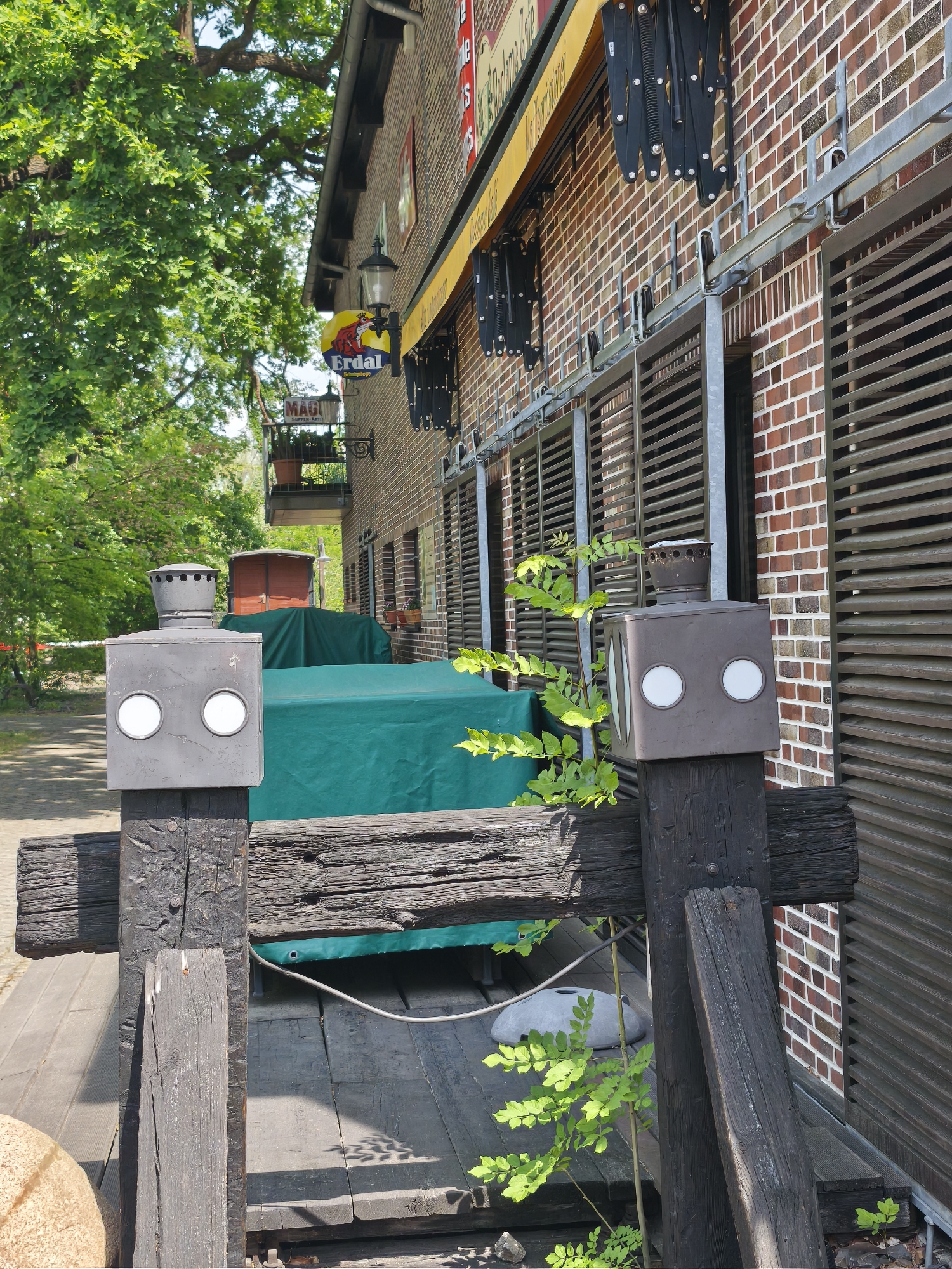 The image depicts the exterior of a brick building with closed, slatted shutters. The location is likely a restaurant or business with outdoor seating, indicated by the green-covered tables and the Erdal sign. In the foreground, there are wooden posts with box-shaped lighting fixtures on top. Some greenery is growing around the posts. The overall scene is located in Unknown, Unknown.