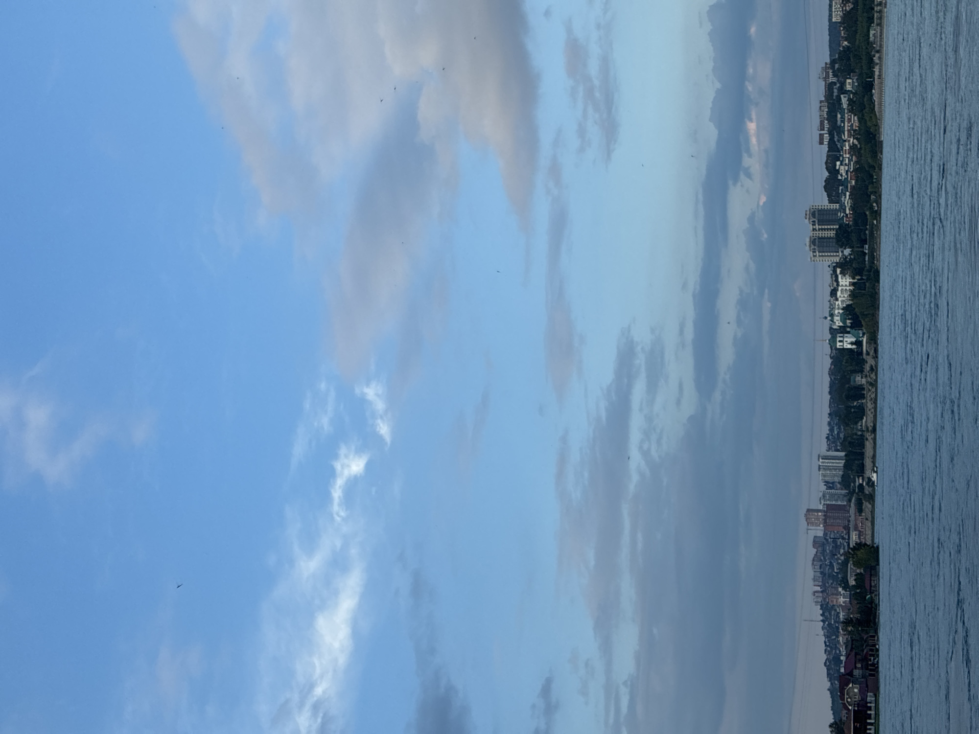The image captures a wide-angle view of a cityscape bordering a body of water, likely the Angara River in Irkutsk, Russia. The sky is a prominent feature, displaying a gradient of blue with scattered clouds ranging from light and wispy to darker, more defined formations. Several small, dark specks are visible in the sky, suggesting birds or insects. The water in the foreground exhibits a rippled texture, indicating movement.
The middle ground reveals a city skyline characterized by a mix of architectural styles. Buildings vary in height and design, including what appear to be apartment complexes, some with modern, multi-story structures, and others that are smaller and closer to the waterfront. A distinct white building with green domes, possibly a church, stands out. Residential areas with houses featuring red roofs are also present. Dense green foliage, likely trees, forms a natural buffer between the water and the urban development. A single white boat is visible on the water, near the shore. The background shows a hilly terrain extending behind the city, with more buildings dotted across the landscape. The overall lighting suggests it is either early morning or late afternoon, with a soft, diffused light.