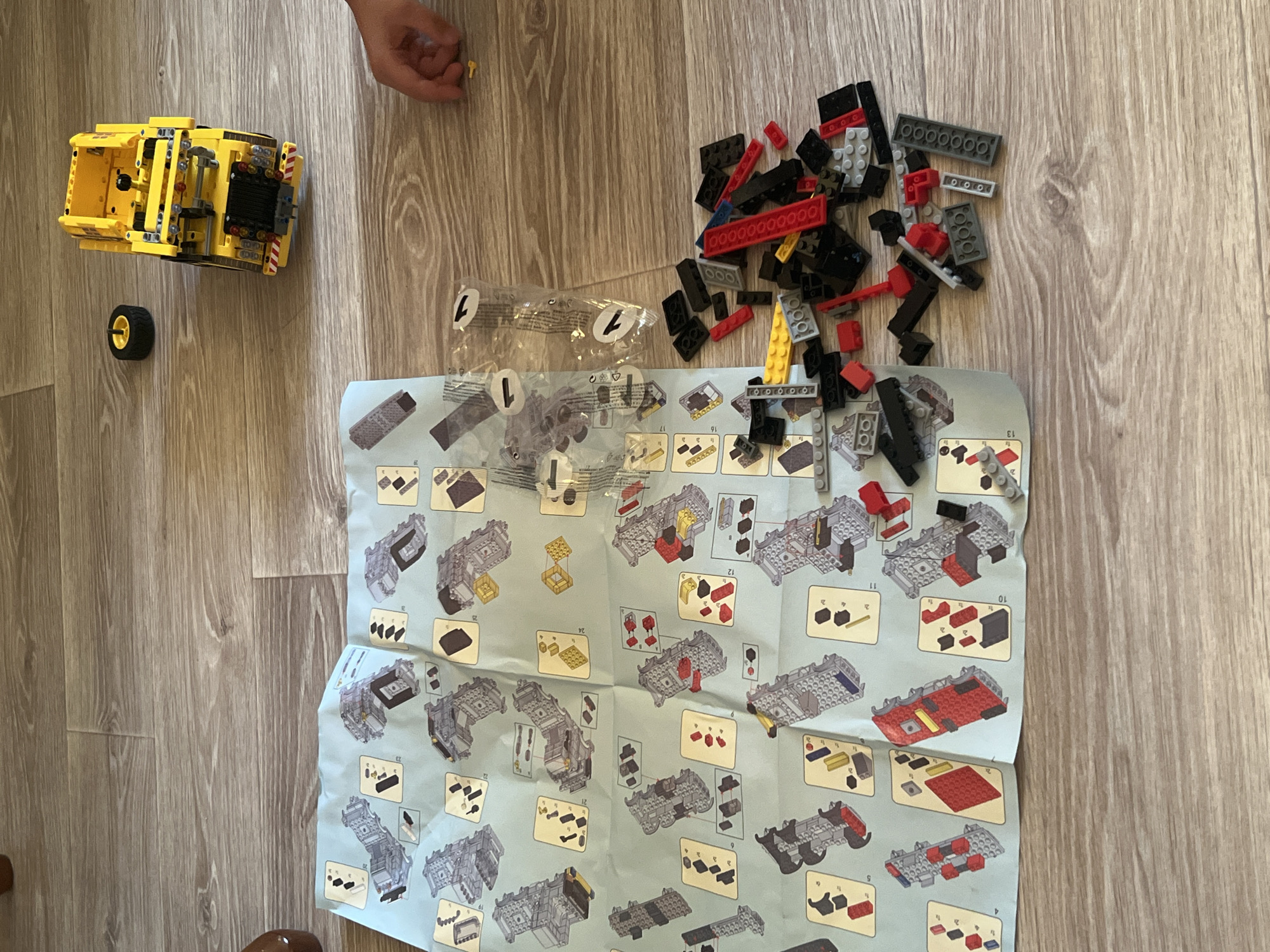 An overhead shot shows a partially assembled yellow LEGO Technic truck, a spare wheel, and instruction manuals spread across a light brown wood-patterned floor. A child's hand is visible in the upper right corner, reaching towards a small yellow LEGO piece on the floor. A pile of assorted red, black, and gray LEGO bricks is scattered to the right of the instruction manuals. The scene suggests an ongoing LEGO building activity. There is no indication of the location being Volgograd, Russia.