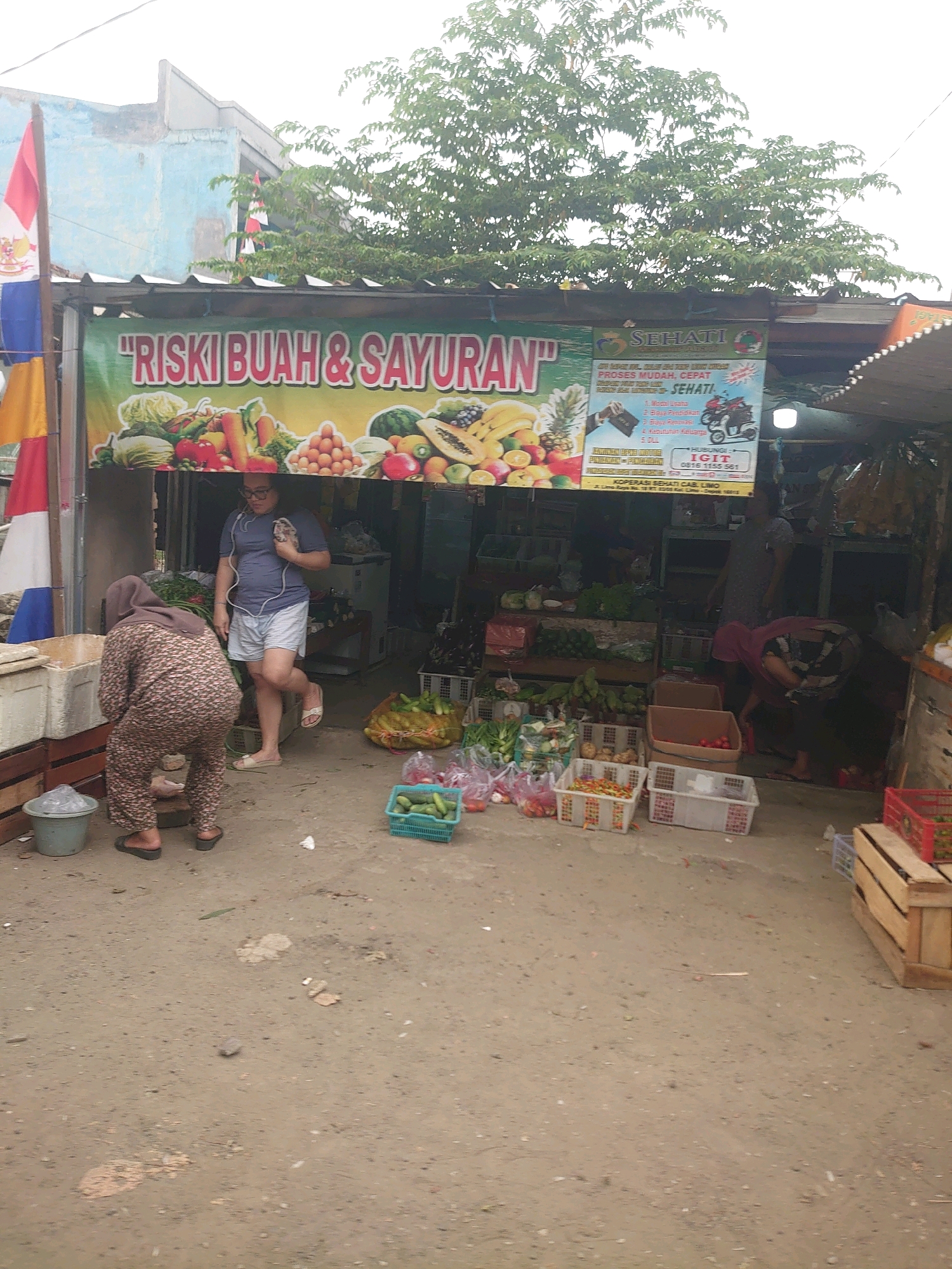 The image depicts a street-level view of an outdoor fruit and vegetable stall in Depok, Indonesia. A banner with the text "RISKI BUAH & SAYURAN" (Riski Fruits & Vegetables) is prominently displayed. To the right of this banner, another sign for "SEHATI" is visible, featuring text related to business and education, along with an image of a motorcycle.
In the foreground, a woman wearing a hijab and a patterned outfit is bent over, appearing to be tending to something near a plastic container. A man wearing glasses, a blue t-shirt, and shorts is walking away from the stall, carrying a small item in his hand. The stall itself is laden with various fresh produce, including vegetables like cucumbers, tomatoes, and leafy greens, as well as fruits such as bananas, watermelons, and pineapples. Several crates and baskets are used to display the goods.
The ground is dirt and gravel, typical of an outdoor market setting. In the background, a building with a light blue facade is visible, and lush green trees overhang the stall. The overall atmosphere suggests a bustling local market environment.